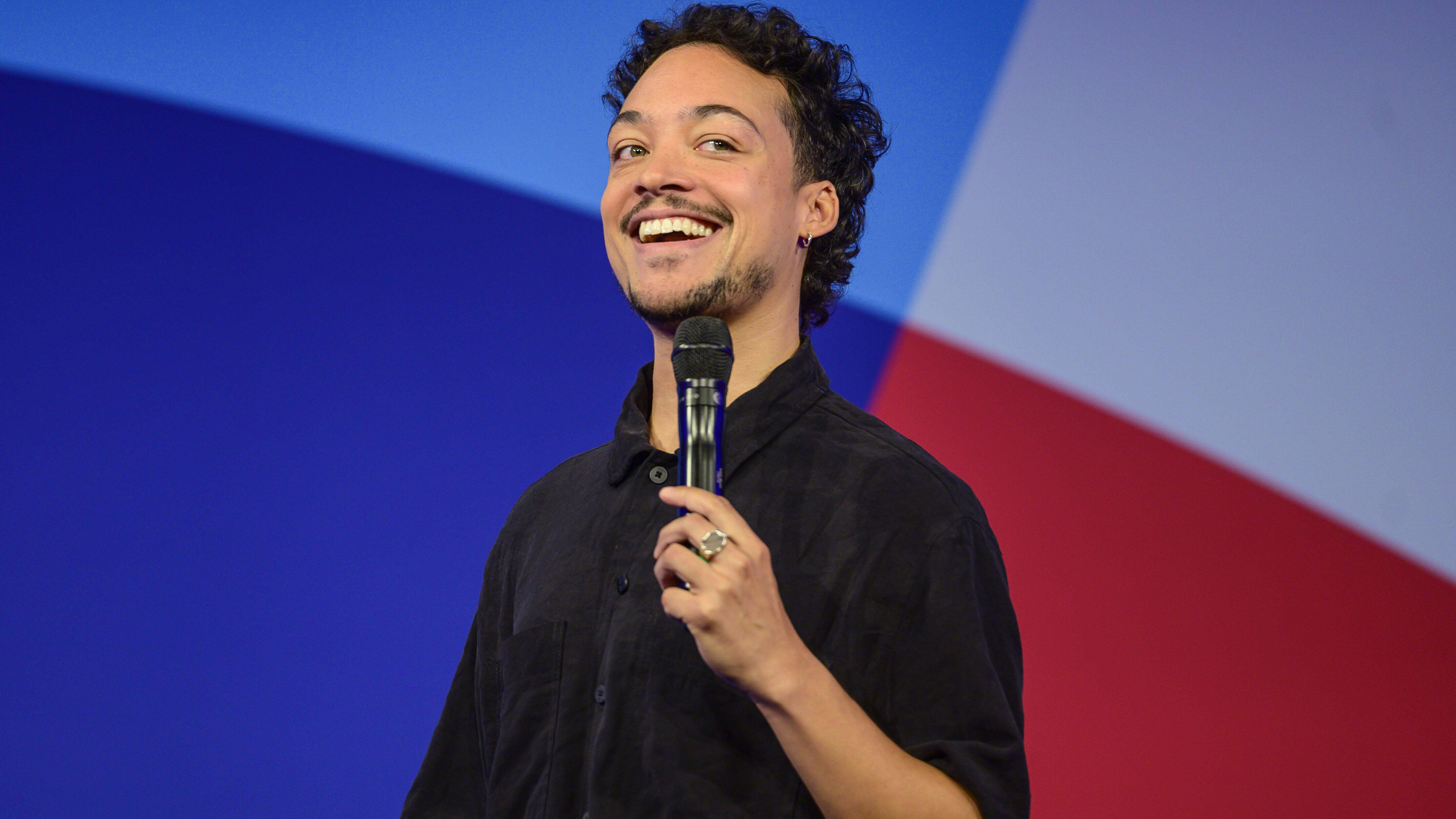 Comedian und Moderator Aurel Mertz steht bei einer Veranstaltung auf der Bühne und hält ein Mikrofon in der Hand.