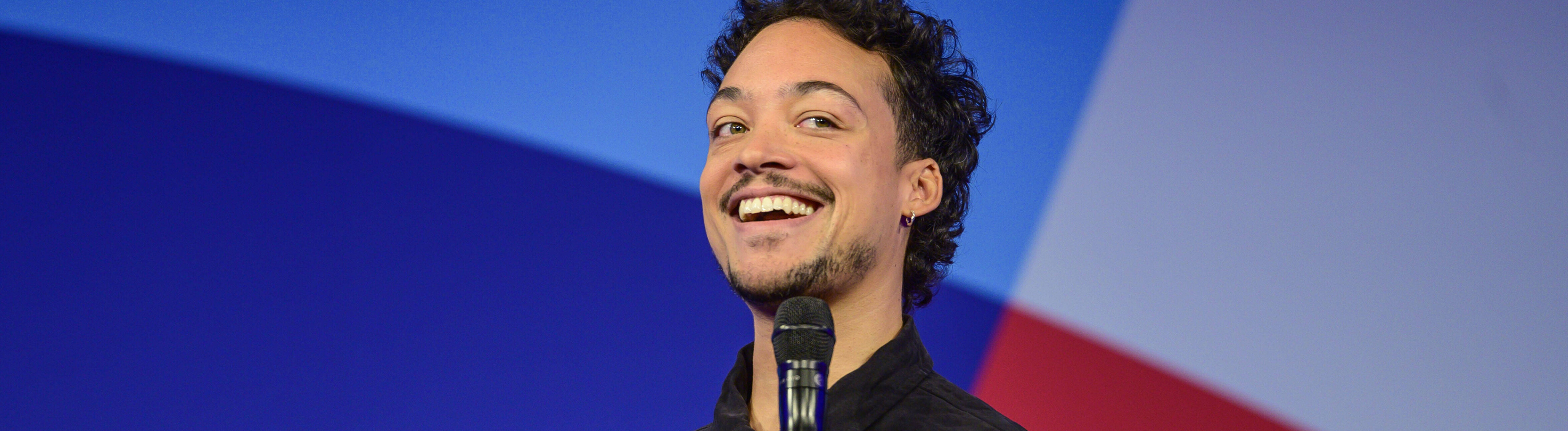Comedian und Moderator Aurel Mertz steht bei einer Veranstaltung auf der Bühne und hält ein Mikrofon in der Hand.