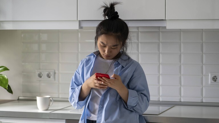 Eine junge Frau steht in einer Küche. Sie lehnt an der Küchenzeile an und schaut auf ihr Handy. Sie wirkt verärgert.