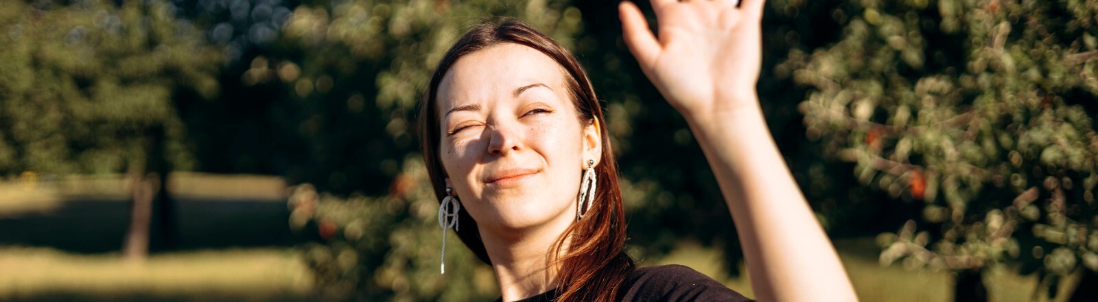 Eine Frau ist an einem sonnigen Tag draußen unterwegs. Sie hält einen orangefarbenen Thermobecher in der einen Hand und die andere Hand vor ihr Gesicht, um sich vor der Sonne zu schützen.