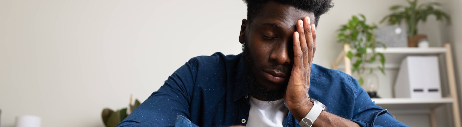Ein junger Mann sitzt auf einer Couch und hält seine Hand an seinen Kopf. Er wirkt müde.