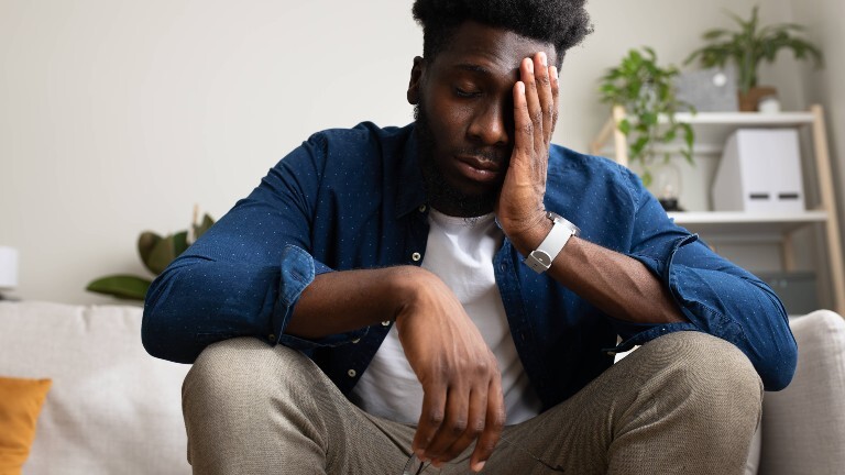 Ein junger Mann sitzt auf einer Couch und hält seine Hand an seinen Kopf. Er wirkt müde.