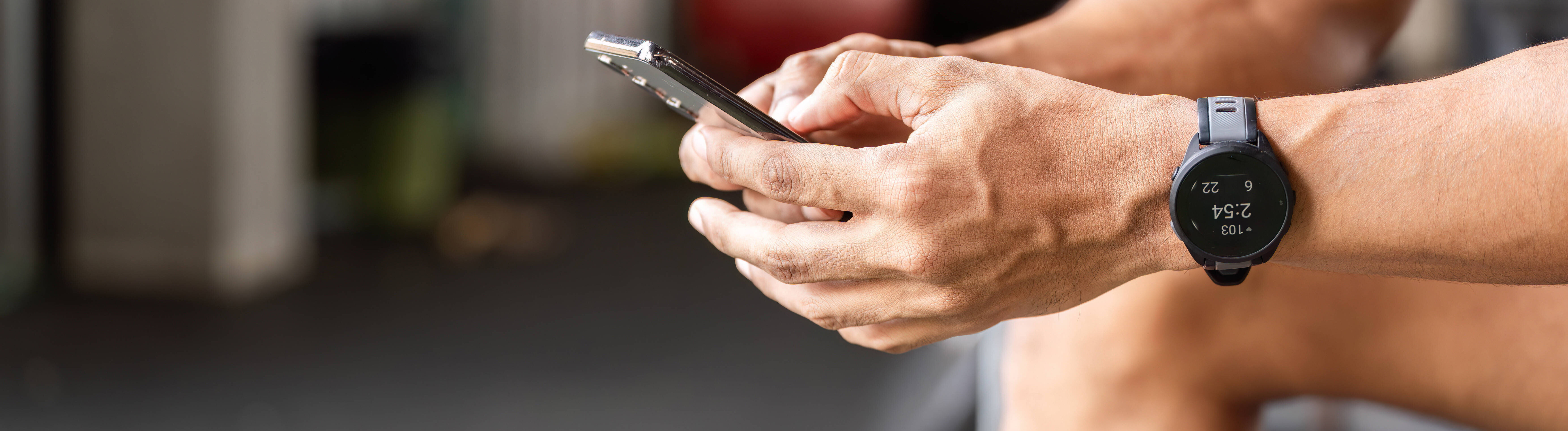 Ein Sportler nutzt eine Fitness-App auf seinem Smartphone während einer Pause im Fitnessstudio. Er sitzt dort, konzentriert auf sein Smartphone und verfolgt mithilfe einer Fitness-App seine Fortschritte und Trainingseinheiten.