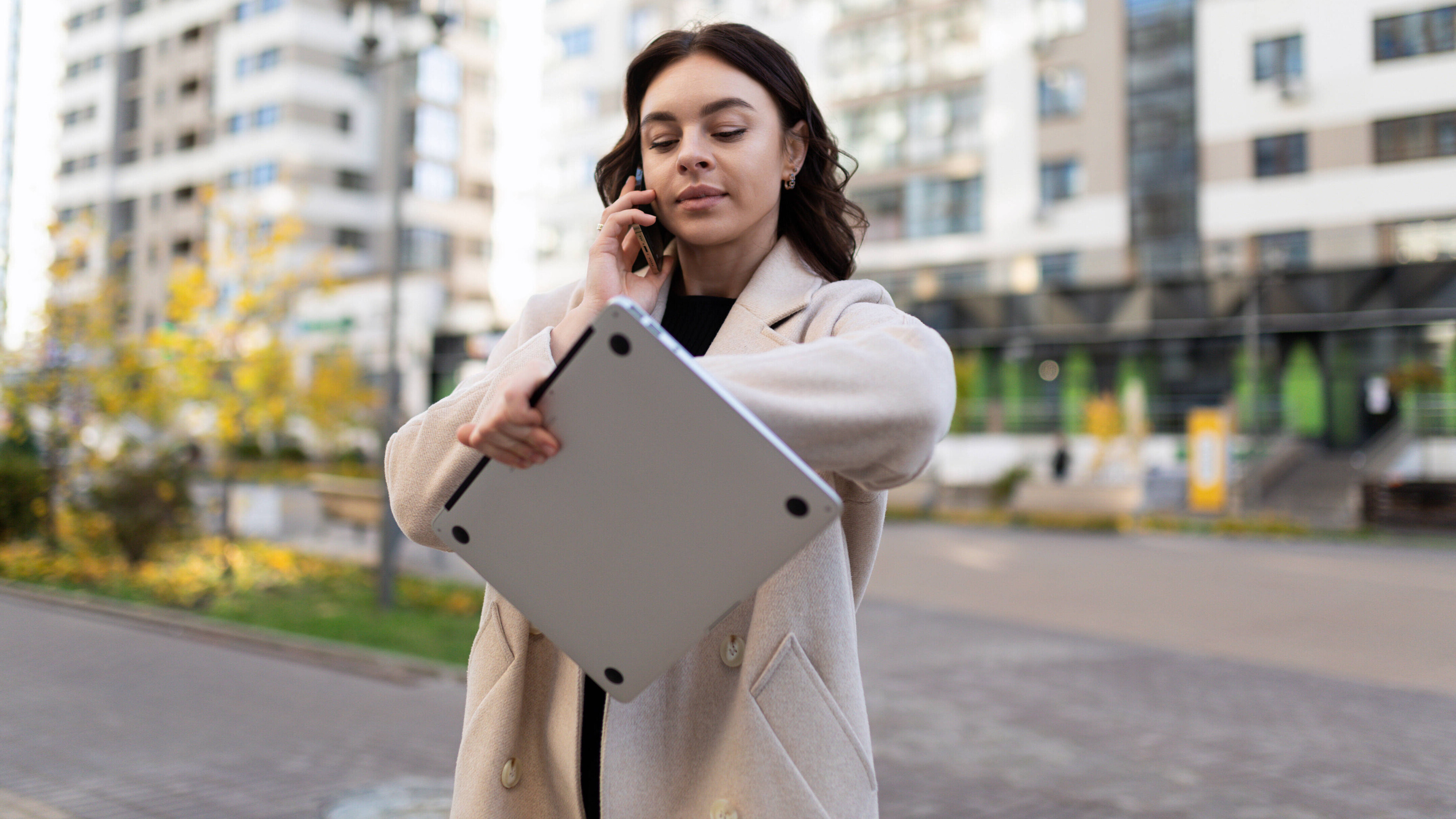 Eine Frau trägt einen Laptop, telefoniert und schaut auf die Uhr
