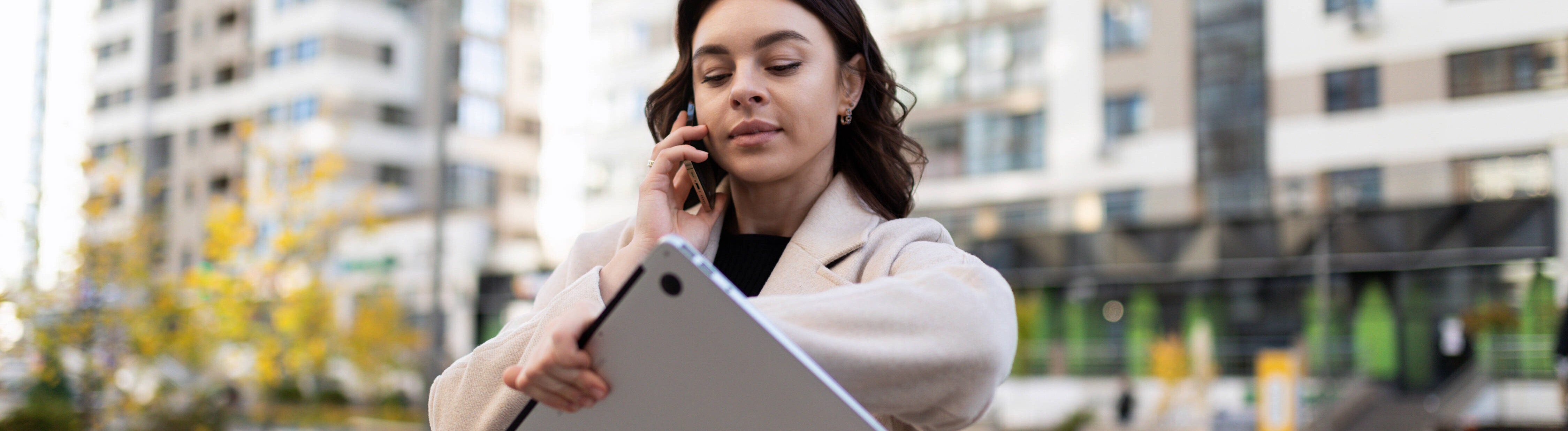 Eine Frau trägt einen Laptop, telefoniert und schaut auf die Uhr