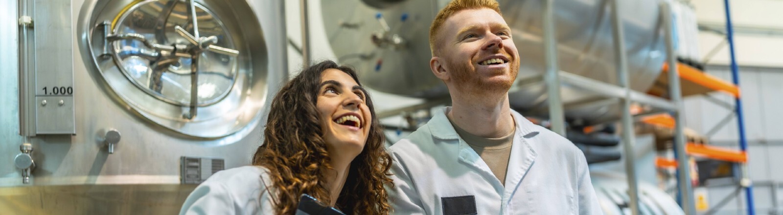 Eine junge Frau und ein junger Mann stehen in Laborkitteln in einer Produktionshalle. Sie halten jeweils Klemmbretter in den Händen und lachen.