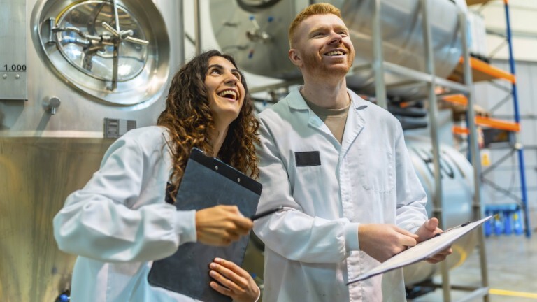 Eine junge Frau und ein junger Mann stehen in Laborkitteln in einer Produktionshalle. Sie halten jeweils Klemmbretter in den Händen und lachen.
