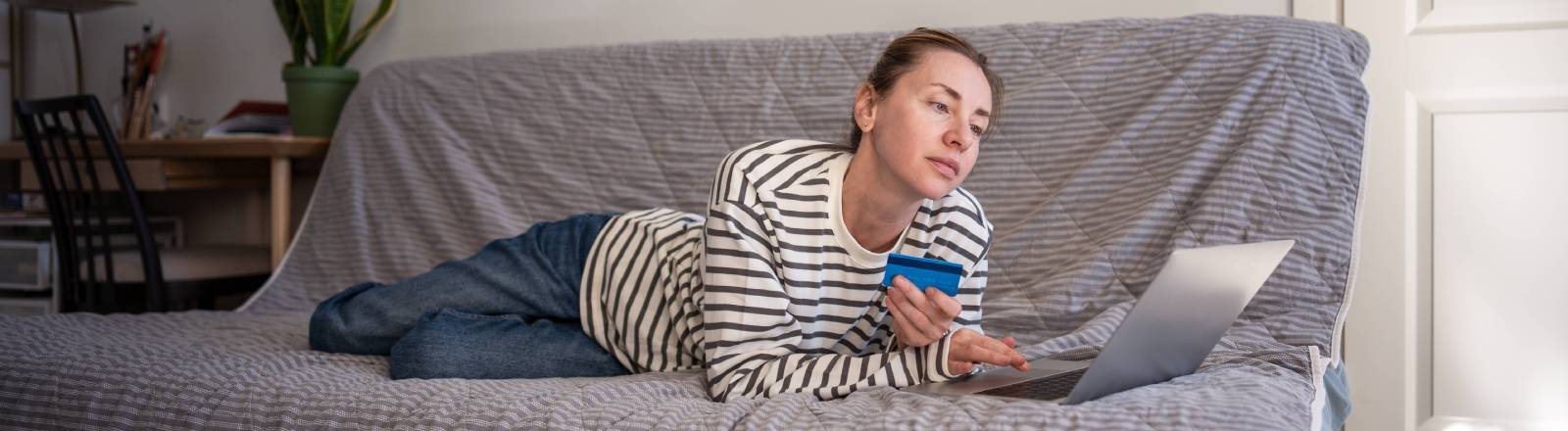 Eine Frau, die mit Bankkarte in der Hand auf der Couch liegt und einen Laptop benutzt
