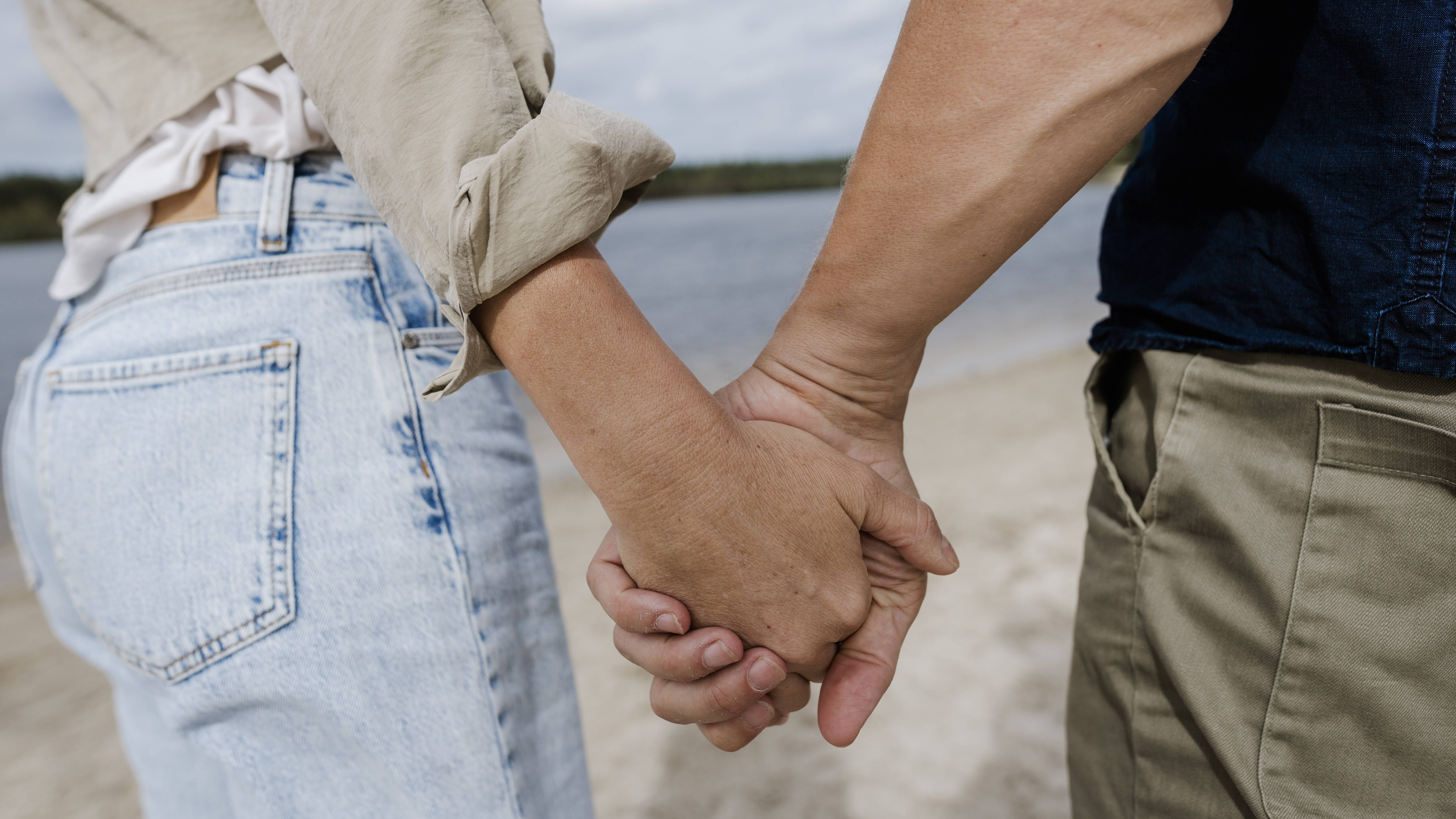 Nahaufnahme der Hände einer jungen Frau und eines jungen Mannes, die am Strand eines Sees im Sonnenschein Händchen halten