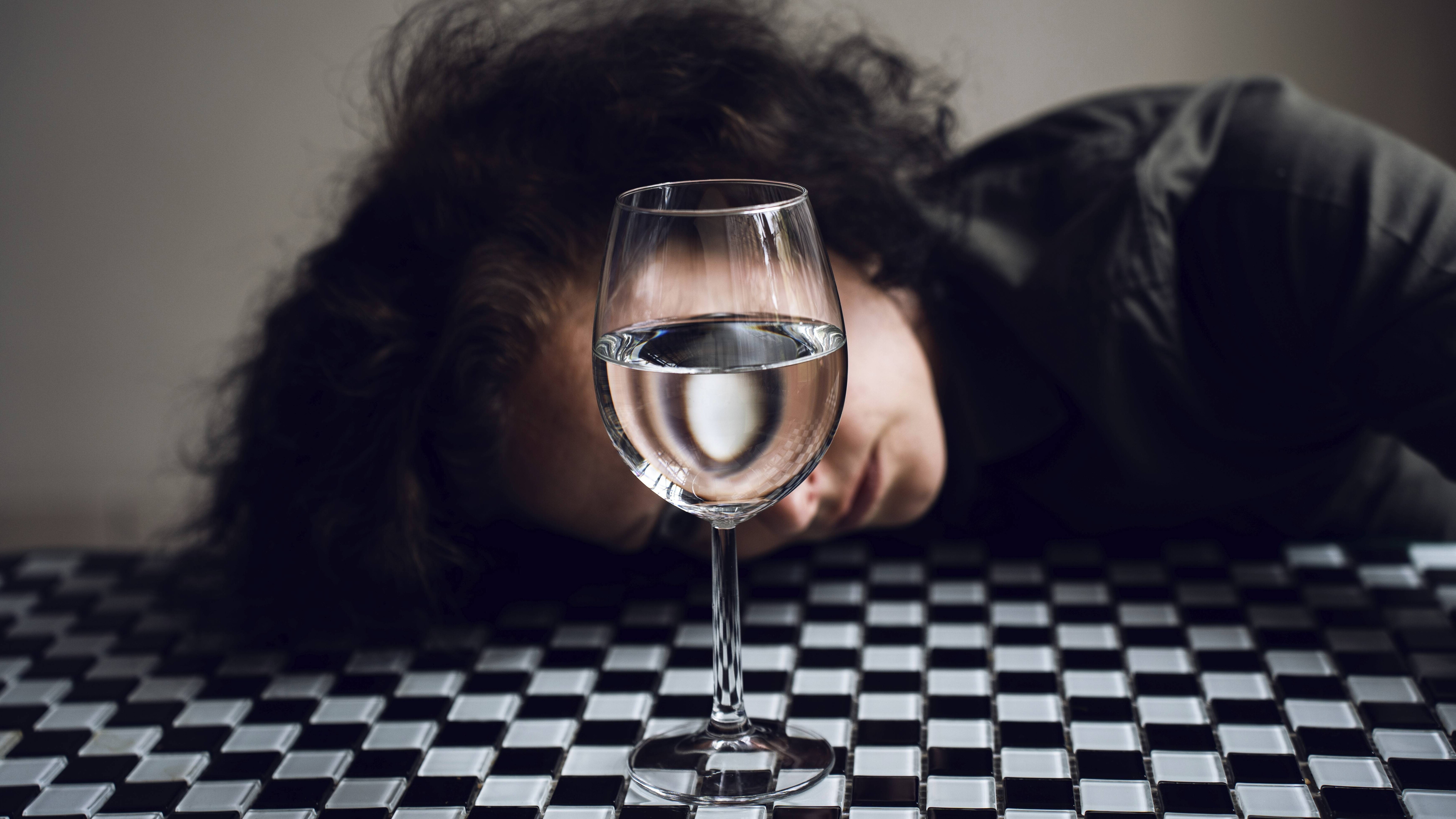 Eine junge Frau hat den Kopf auf einen schachbrettgemusterten Tisch gelegt, ihr Gesicht ist von einem mit Wasser gefüllten Weinglas verdeckt.
