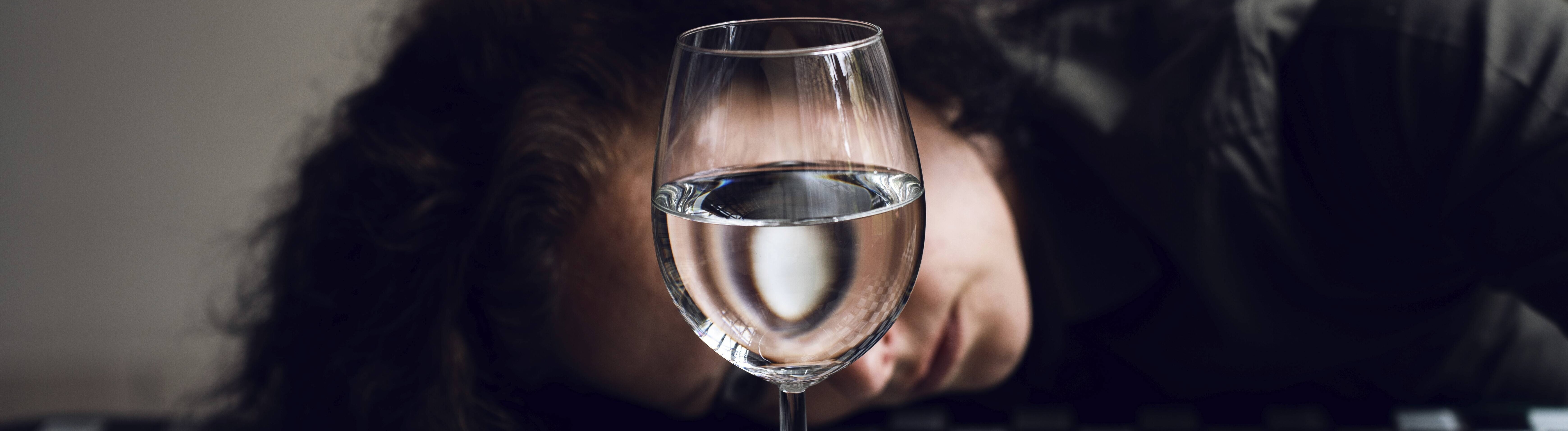 Eine junge Frau hat den Kopf auf einen schachbrettgemusterten Tisch gelegt, ihr Gesicht ist von einem mit Wasser gefüllten Weinglas verdeckt.