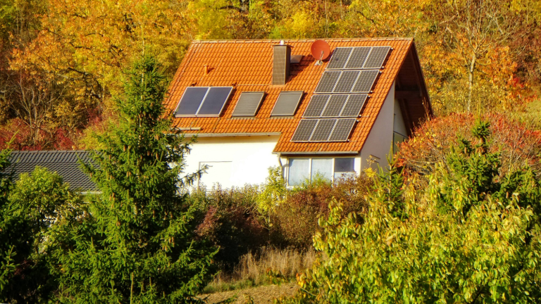 Inmitten eines herbstlich aussehenden Waldes steht ein Einfamilienhaus. Auf dem Dach befinden sich Solaranlagen.