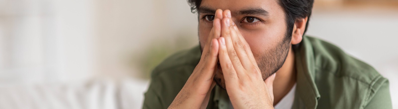 Ein Mann sitzt auf einer Couch und hält sich seine Hände vor das Gesicht, dadurch wird seine Nase verdeckt.