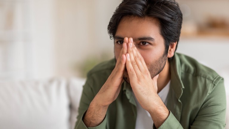 Ein Mann sitzt auf einer Couch und hält sich seine Hände vor das Gesicht, dadurch wird seine Nase verdeckt.