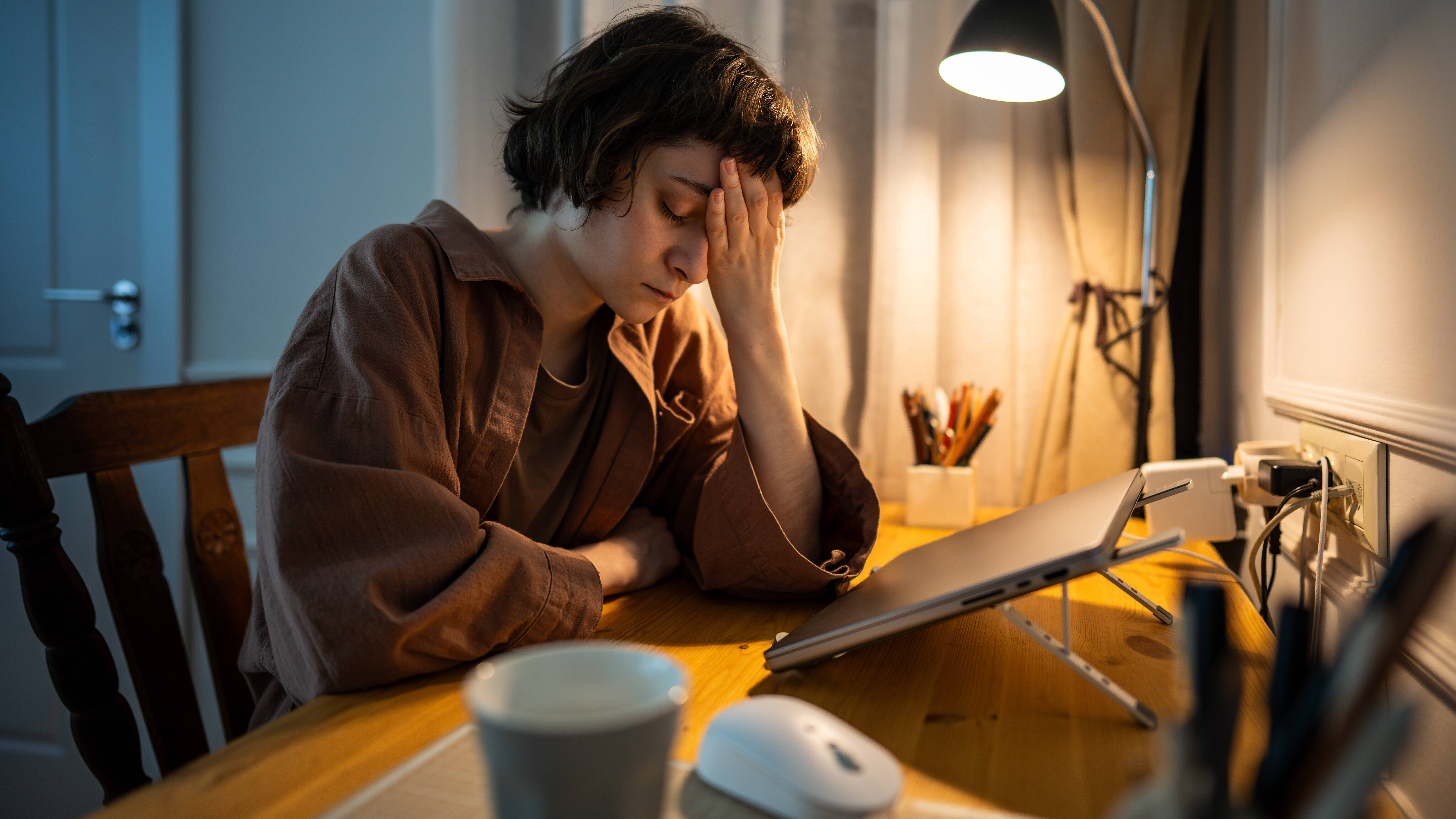 Eine junge Frau sitzt übermüdet an einem Tisch. Vor ihr steht ein Laptop eine Maus und eine Kaffeetasse.