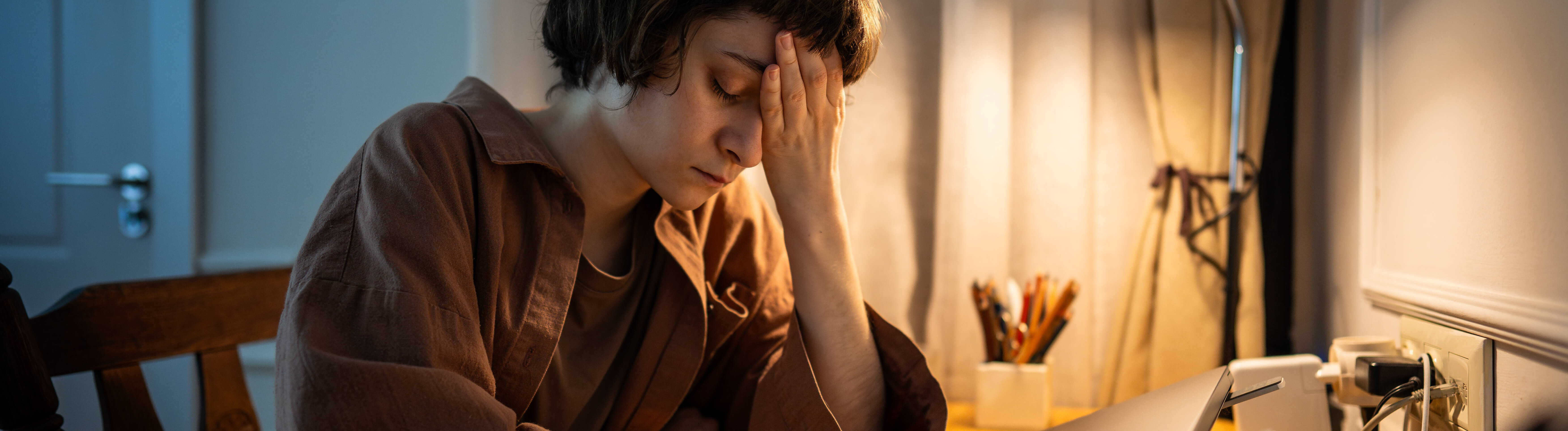 Eine junge Frau sitzt übermüdet an einem Tisch. Vor ihr steht ein Laptop eine Maus und eine Kaffeetasse.