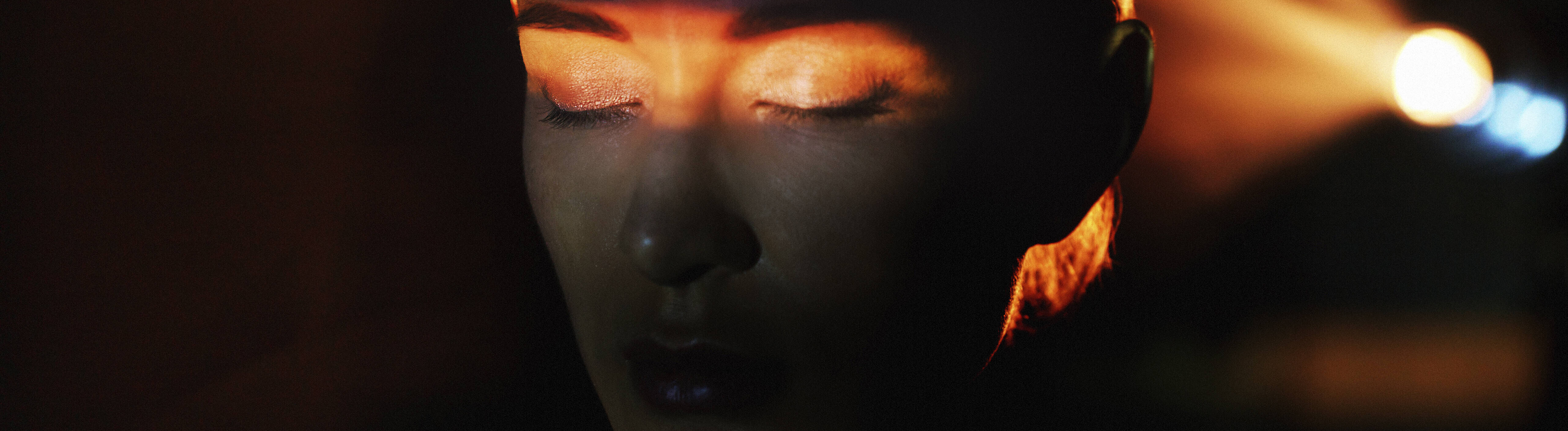 Eine Frau ist alleine in einem abgedunkelten Raum. Hinter ihr leuchtet ein Licht. Sie hat ihre Augen geschlossen. Auf ihre Augen leuchtet ein Lichtstrahl.