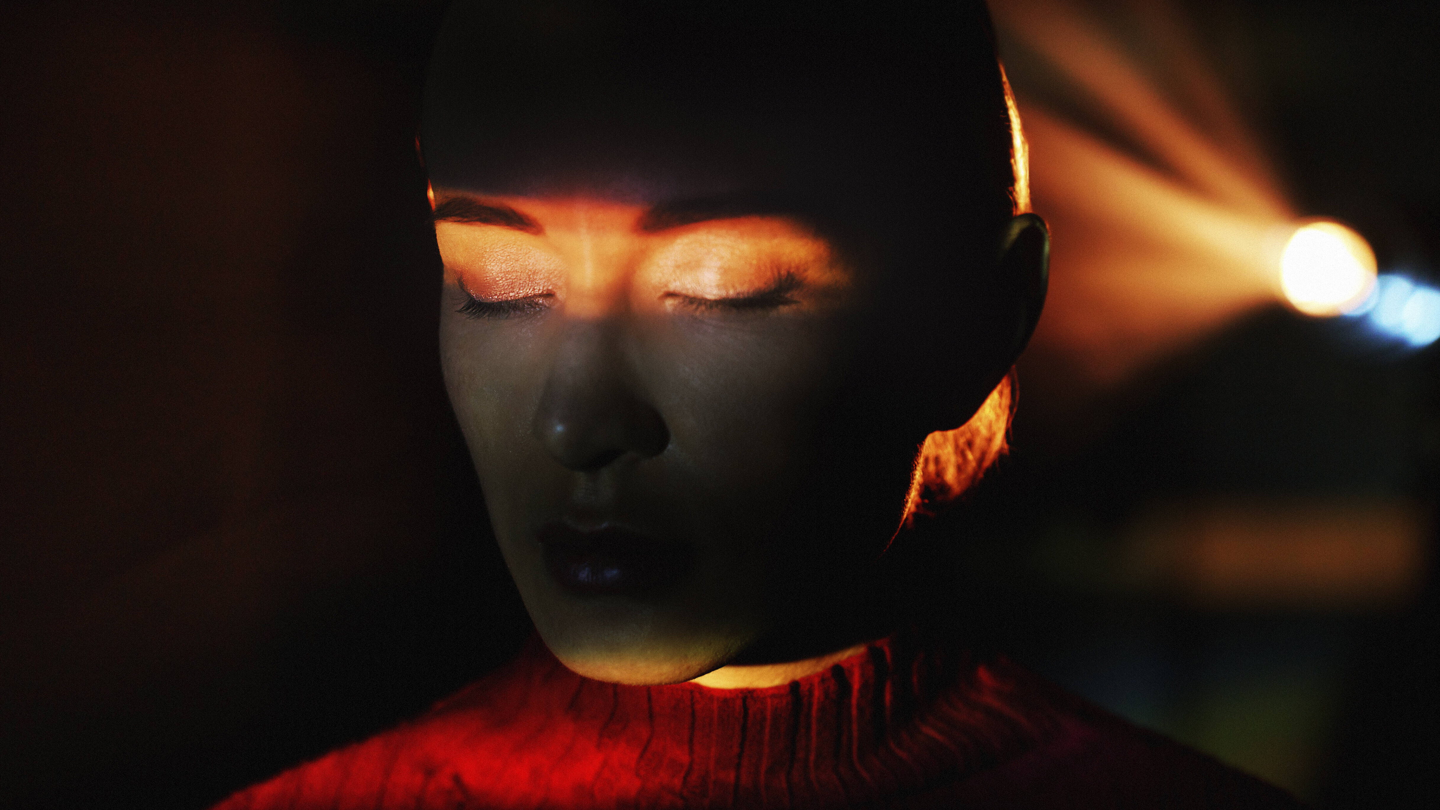 Eine Frau ist alleine in einem abgedunkelten Raum. Hinter ihr leuchtet ein Licht. Sie hat ihre Augen geschlossen. Auf ihre Augen leuchtet ein Lichtstrahl.