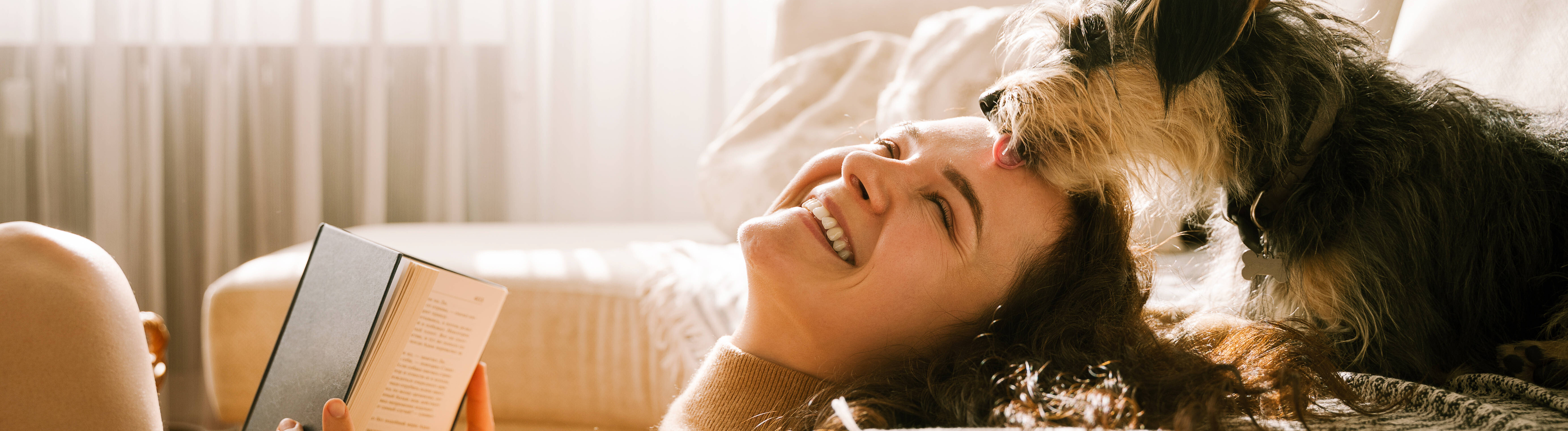 Eine Frau sitzt in einem weiß eingerichteten Zimmer auf dem Boden und lehnt ihren Rücken an einer Couch an. Dort sitzt ein Hund, der die Frau am Kopf berührt.