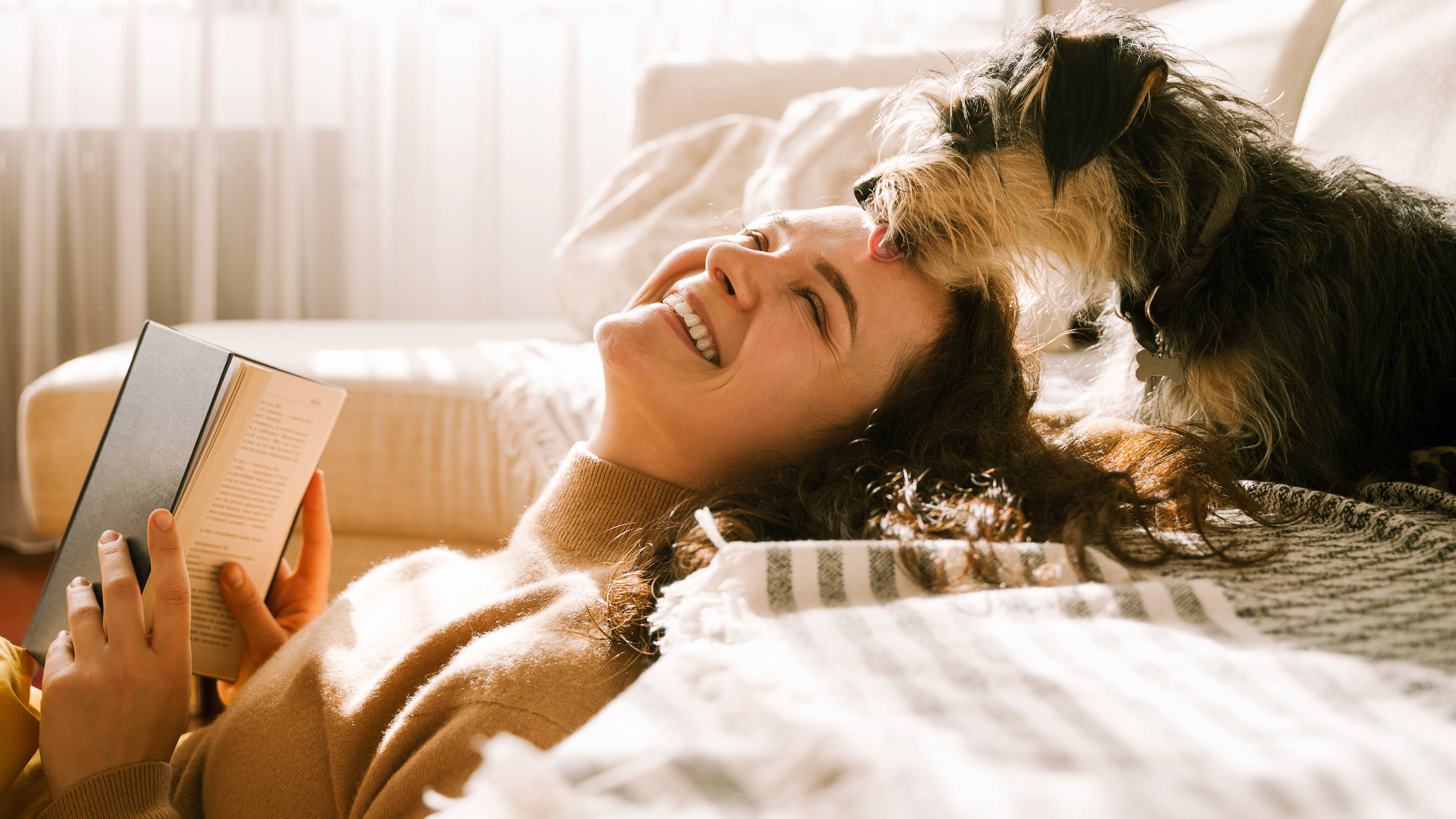 Eine Frau sitzt in einem weiß eingerichteten Zimmer auf dem Boden und lehnt ihren Rücken an einer Couch an. Dort sitzt ein Hund, der die Frau am Kopf berührt.