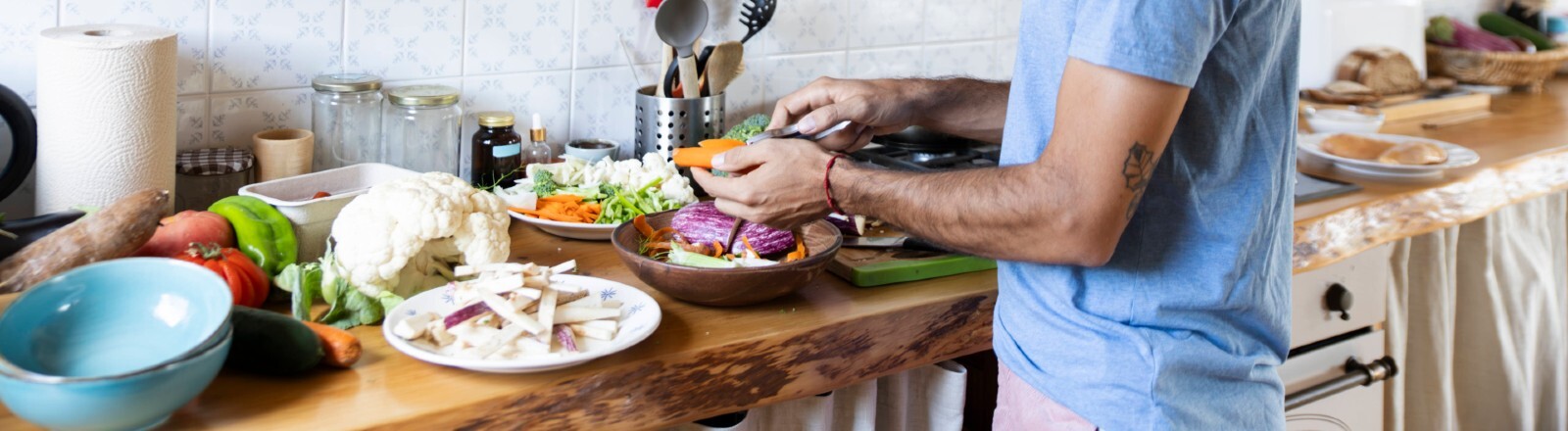 Ein Mann steht lächelnd in einer Küche und bereitet dabei Gemüse zu. Er hat einen kabellosen Kopfhörer im Ohr.