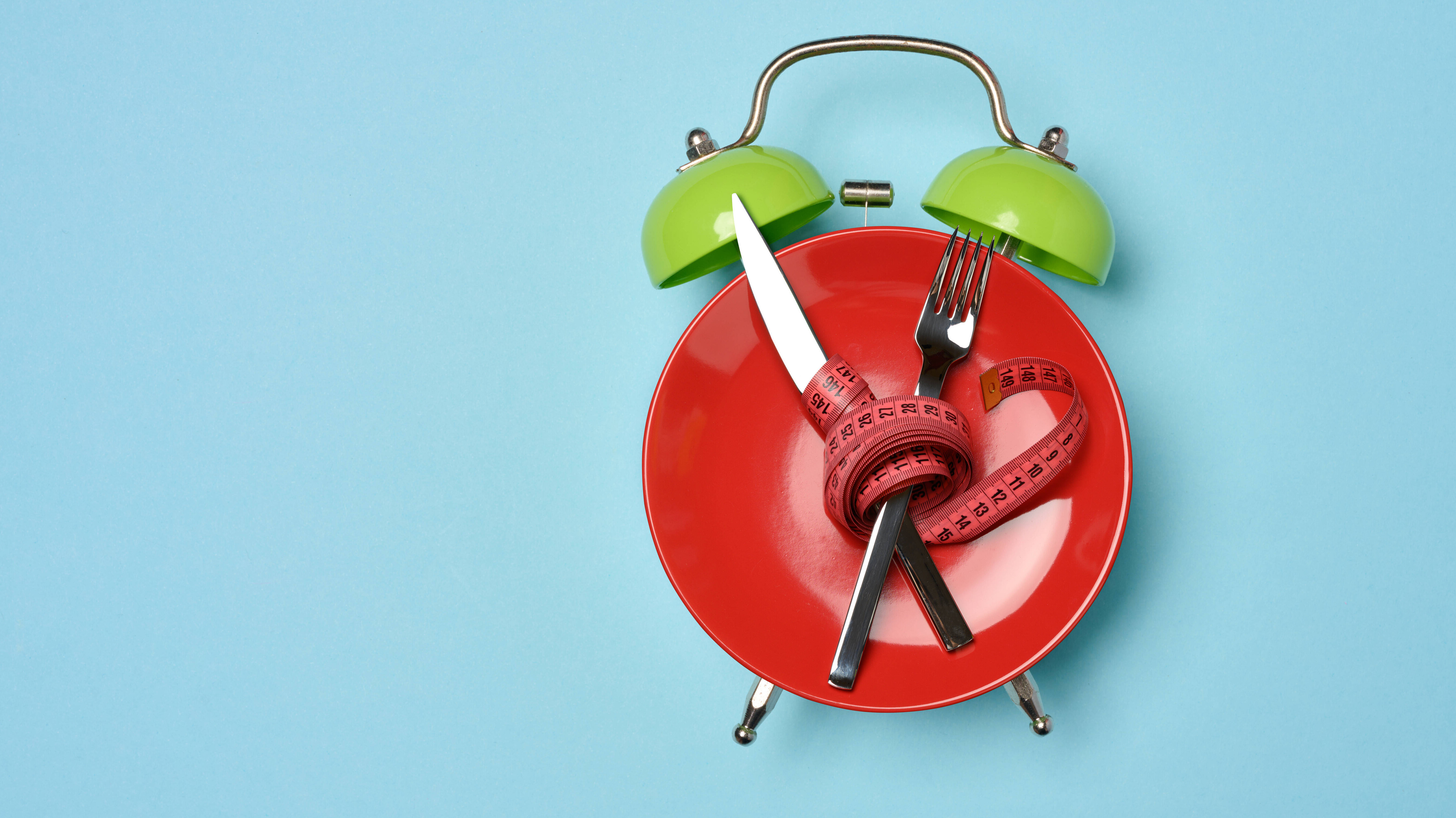 Diät-Konzeptbild, roter Teller, der zum Wecker stilisiert wurde, mit gekreuztem Besteck umwickelt mit rotem Maßband