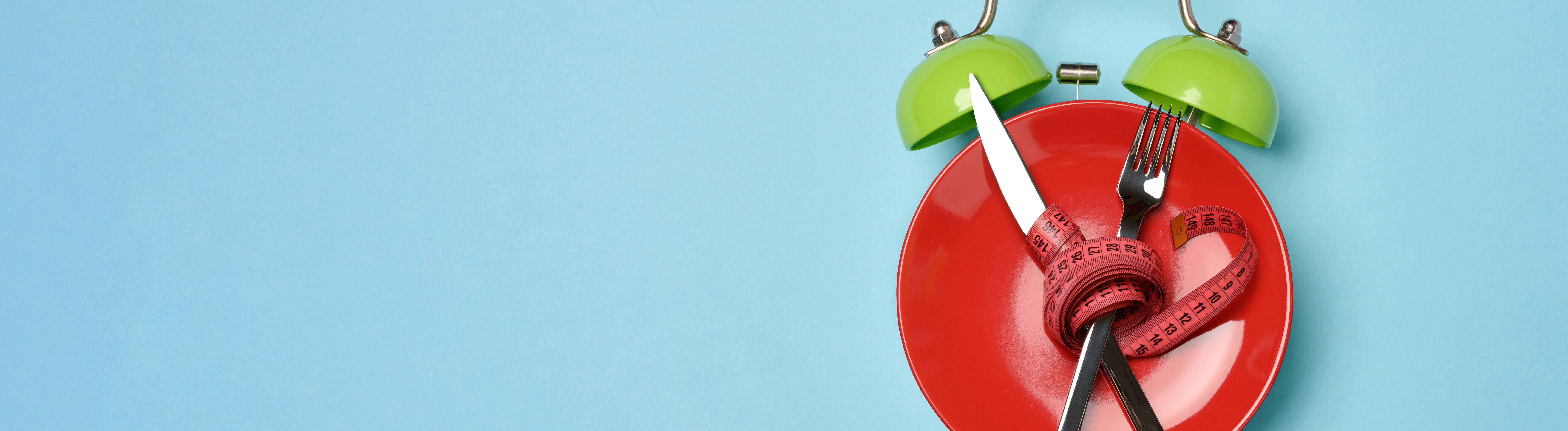 Diät-Konzeptbild, roter Teller, der zum Wecker stilisiert wurde, mit gekreuztem Besteck umwickelt mit rotem Maßband