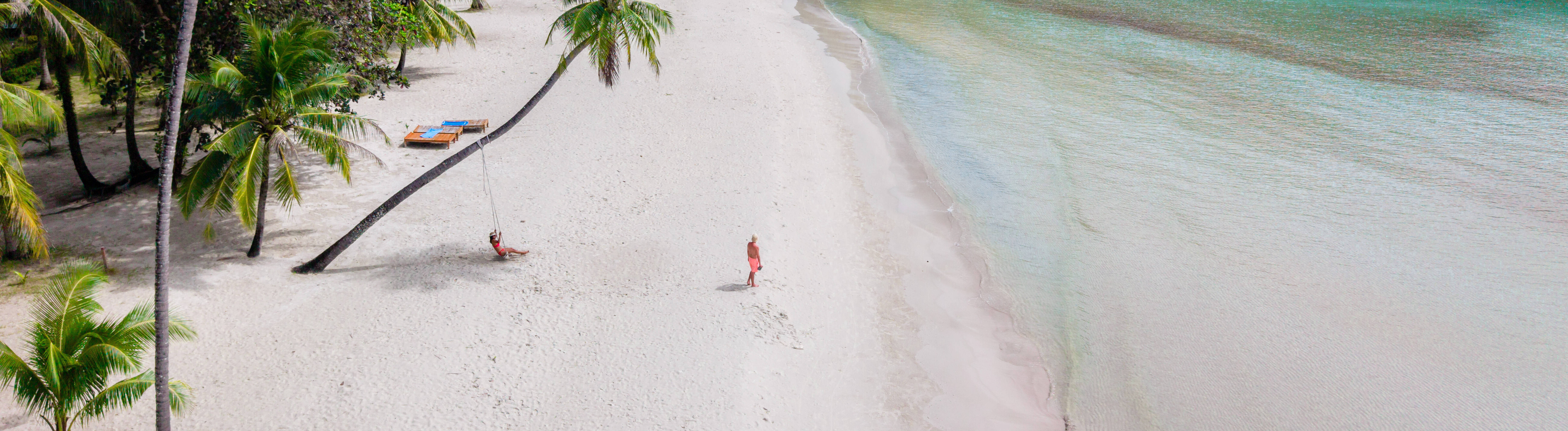 Ein Paar ist an einem Sandstrand umgeben von Palmen und Natur. Die Frau sitzt in einer Schaukel. Der Mann geht Richtung Meer.