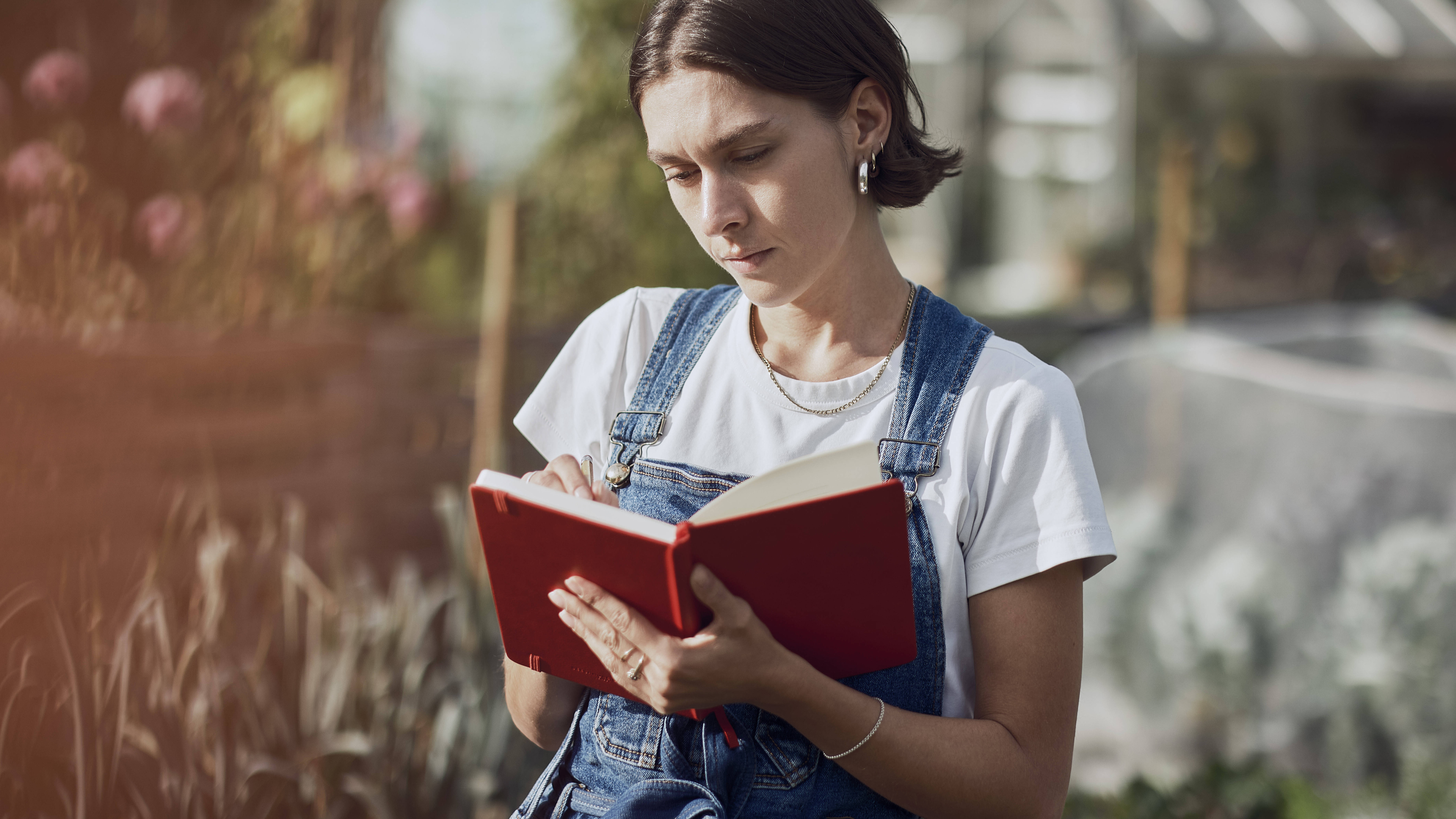 Eine Frau steht in einem Garten und schreibt etwas in ein rotes Notizbuch.