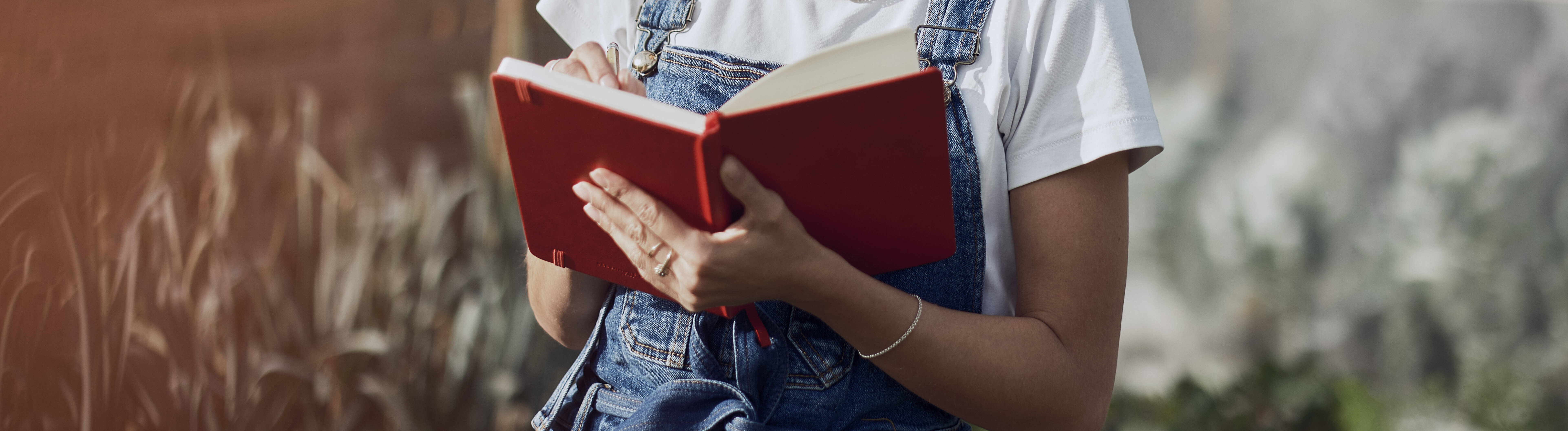 Eine Frau steht in einem Garten und schreibt etwas in ein rotes Notizbuch.