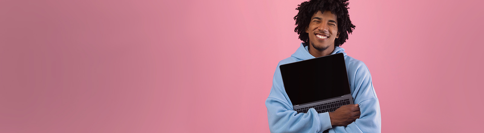 Foto eines jungen Mannes in blauem Pullover vor einer pinken Wand, der ein aufgeklapptes Laptop umarmt