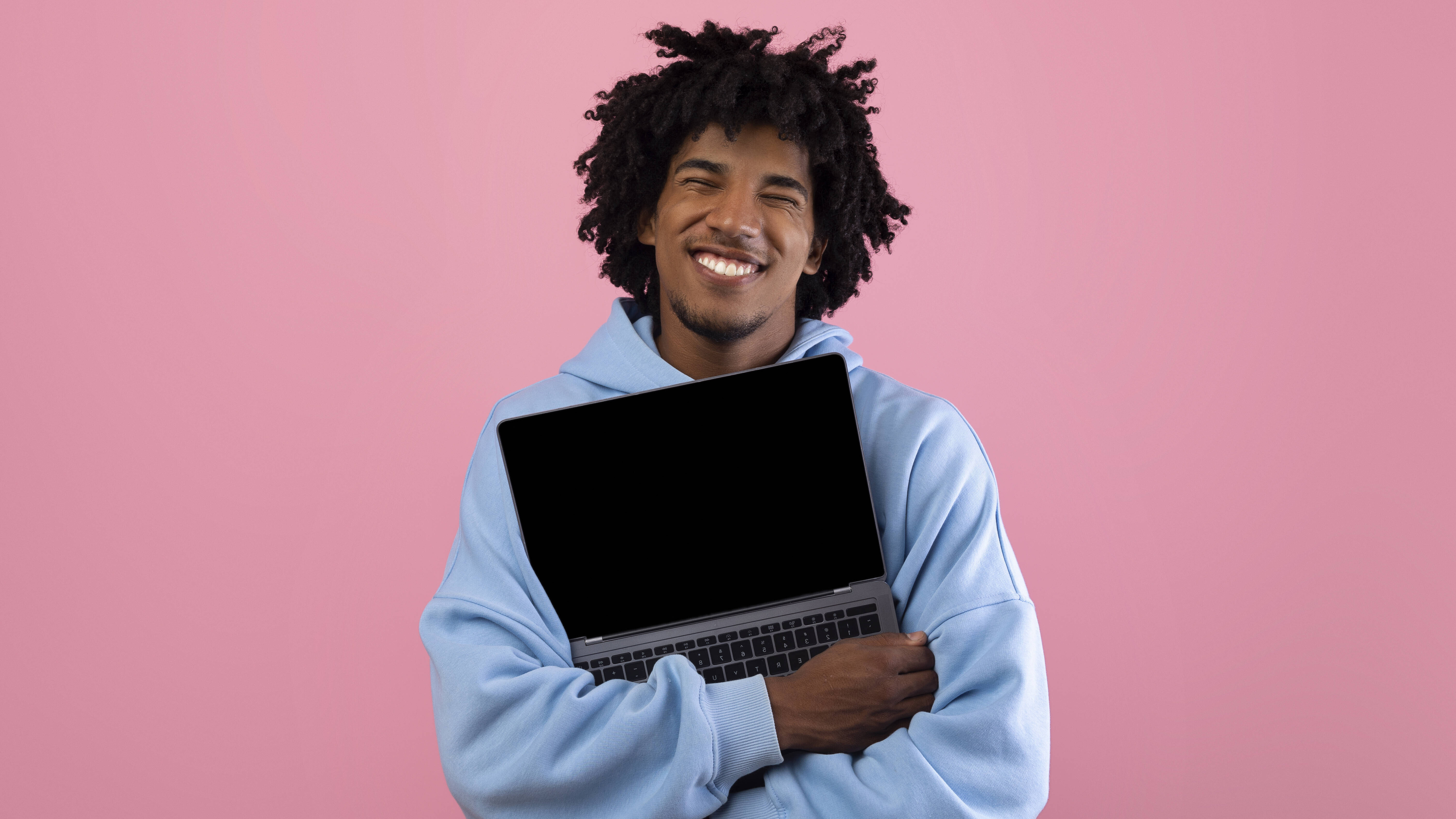 Foto eines jungen Mannes in blauem Pullover vor einer pinken Wand, der ein aufgeklapptes Laptop umarmt