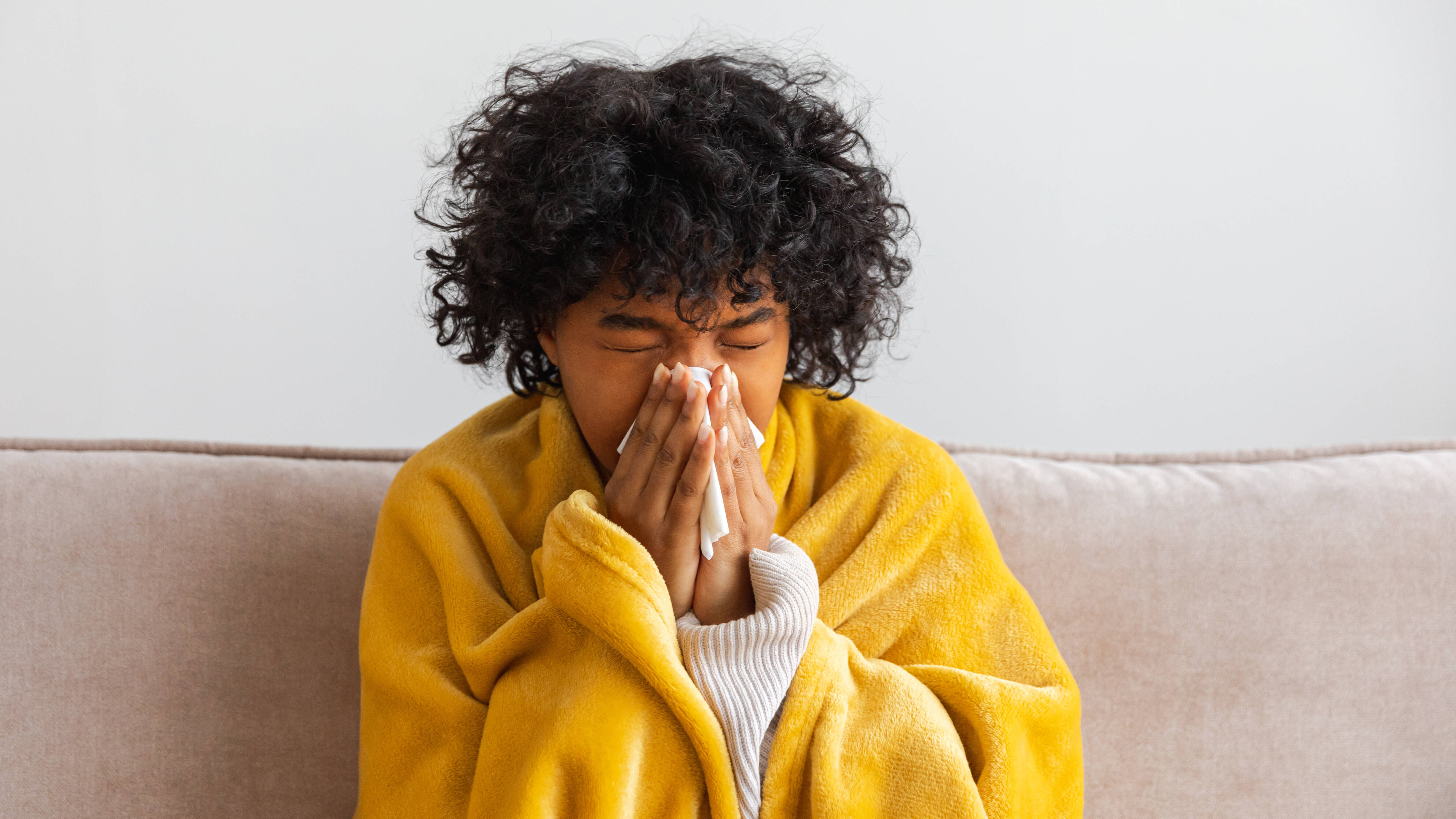 Unglückliche, erkrankte junge Afroamerikanerin mit Erkältung sitzt zu Hause auf dem Sofa. Mädchen bedeckt sich mit einer Decke und putzt sich die Nase mit einem Taschentuch.