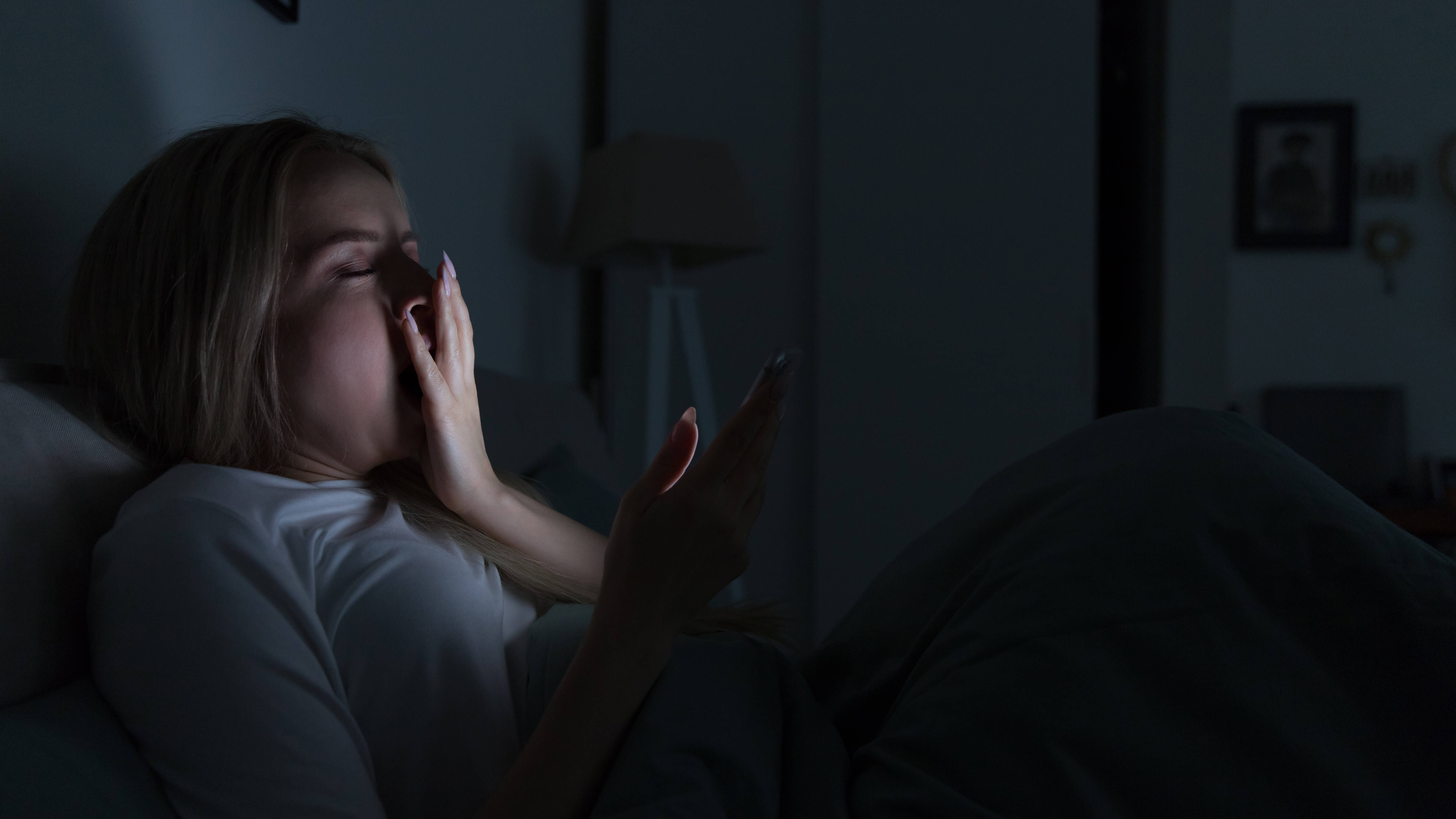 Schläfrige, müde Frau liegt im Bett, benutzt ihr Smartphone, bedeckt ihren Mund mit der Hand und gähnt.