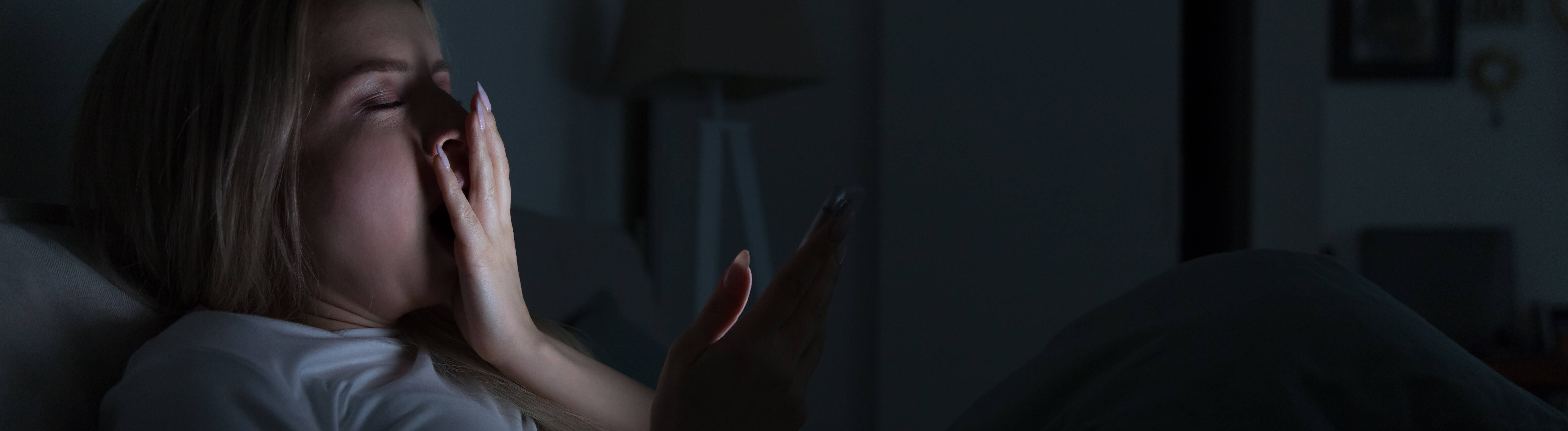 Schläfrige, müde Frau liegt im Bett, benutzt ihr Smartphone, bedeckt ihren Mund mit der Hand und gähnt.