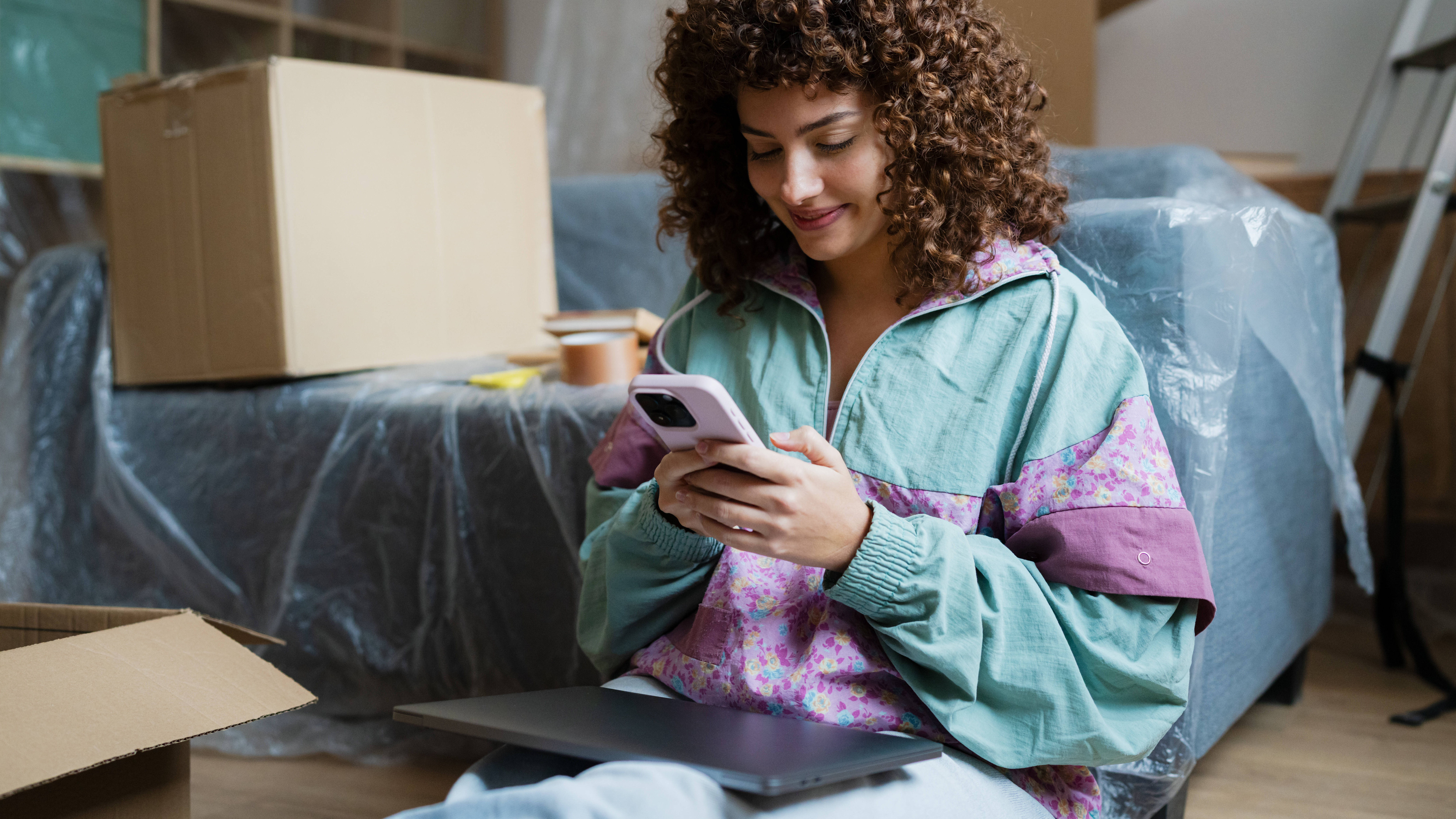 Eine Frau sitzt in einem Wohnzimmer und schaut auf ihr Handy. Um sie herum stehen Umzugskartons. Über den Möbeln liegt eine Plastikfolie.