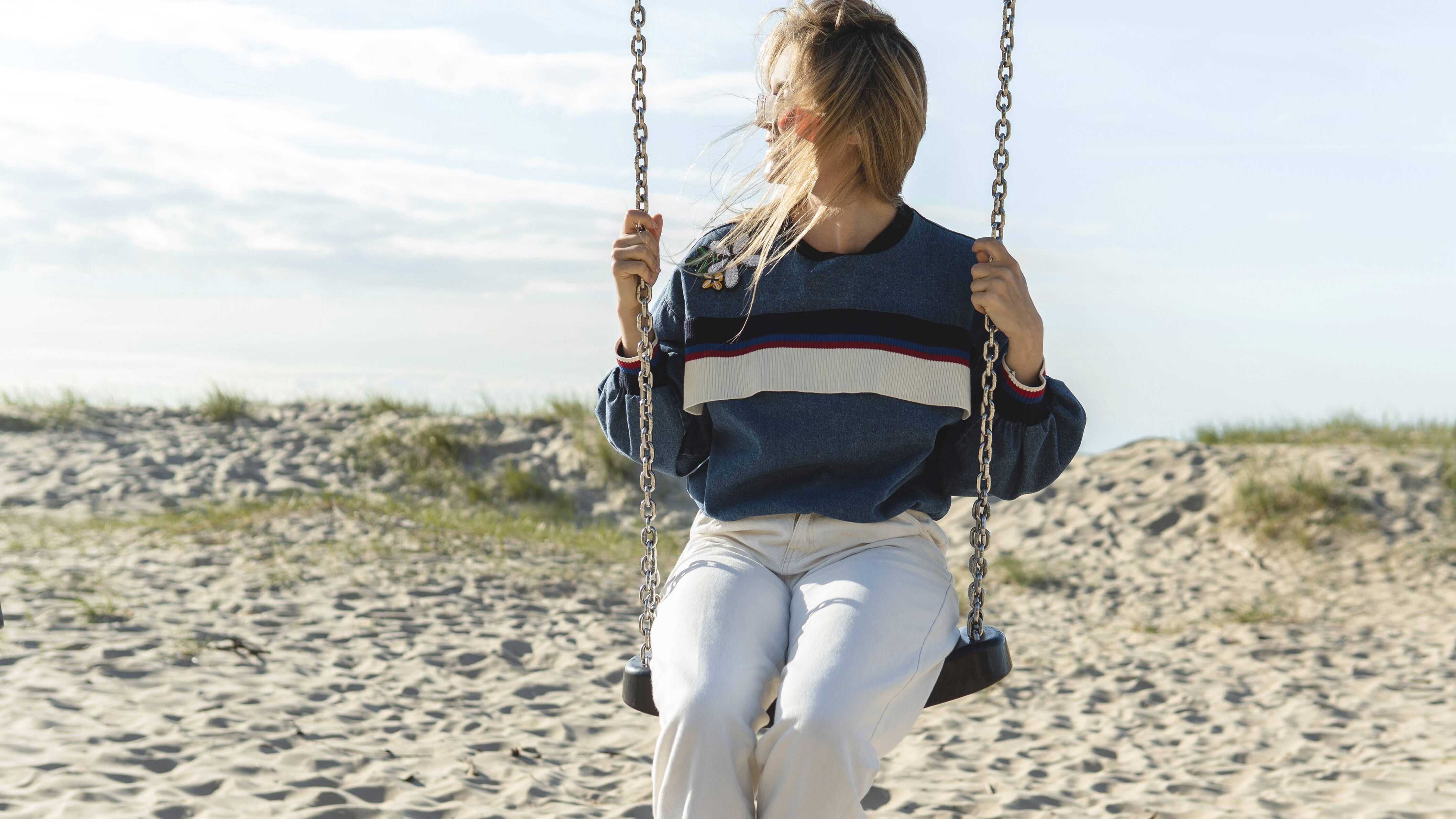 Eine Frau sitzt auf einer Schaukel und schaut zur Seite. Die Schaukel steht an einem Strand. Es scheint die Sonne.