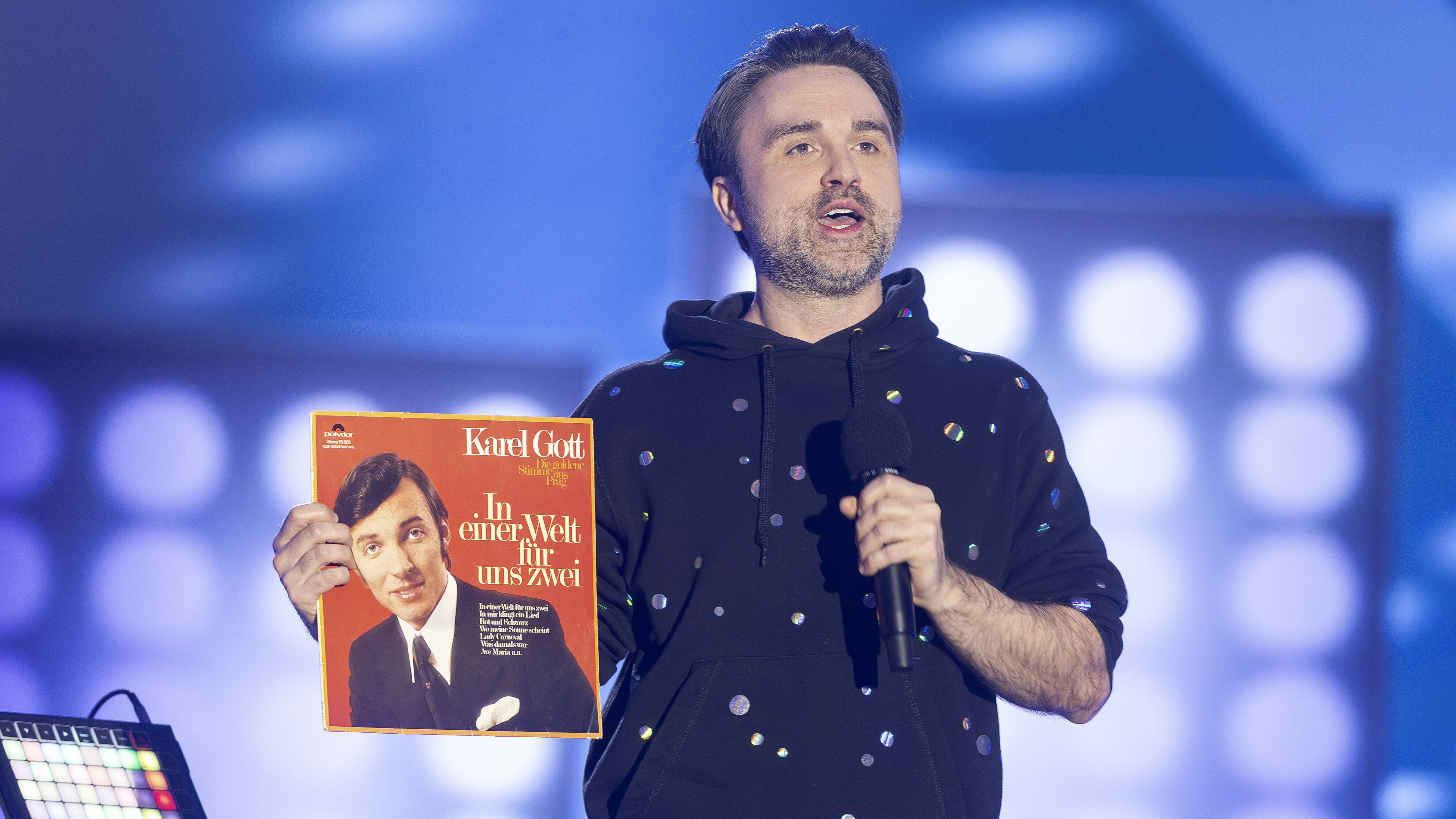 Musikkabarettist und -wissenschaftler Dr. Pop (mit bürgerlichem Namen Markus Henrik) spricht auf der Bühne in ein Mikrofon und hält im rechten Arm eine Schallplatte von Karel Gott (MDR Humorzone, 15.03.2026)