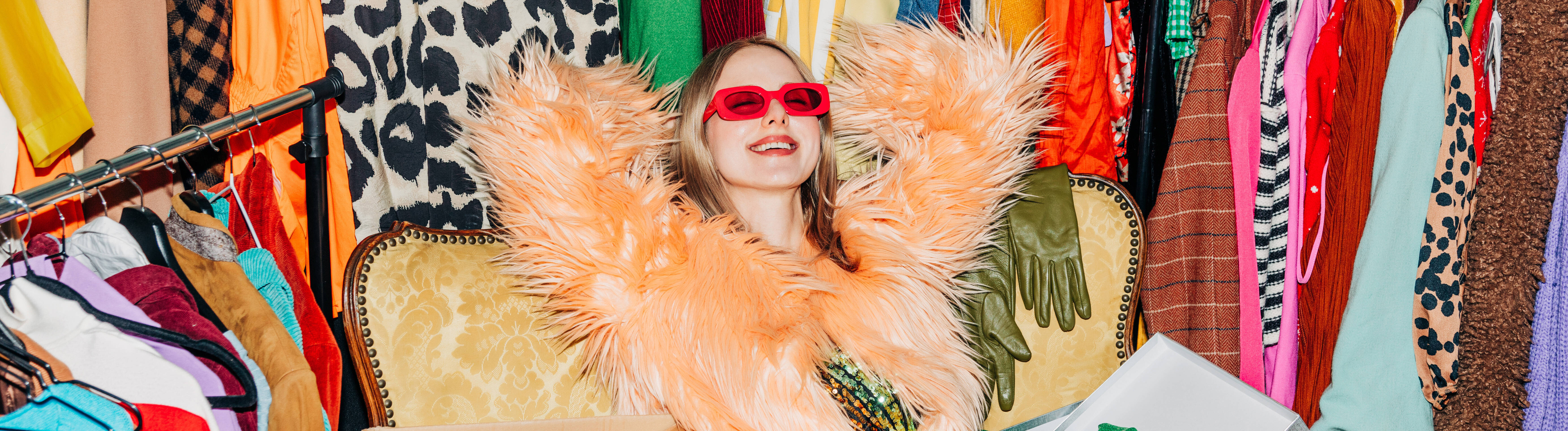 Eine Frau lehnt sich auf einem Sessel zurück. Um sie herum stehen einige Kleiderstangen, die vollgepackt mit Kleidung sind. Die Frau lächelt und trägt eine rote Sonnenbrille.