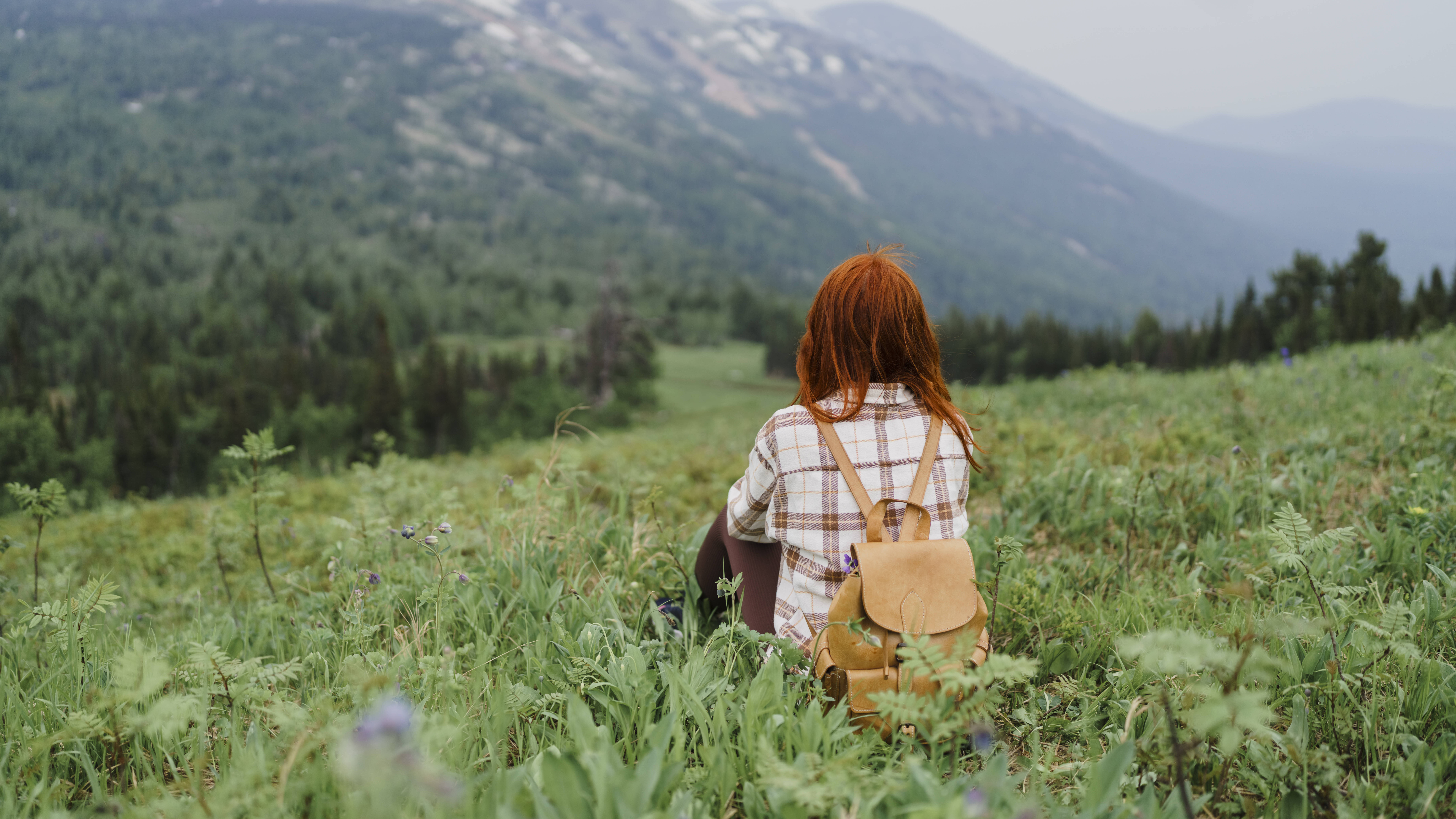 Eine junge Frau mit roten Haaren und Rucksack sitzt im Gras und blickt auf eine weite Berglandschaft.
