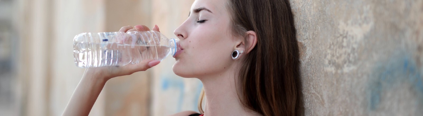 Eine Frau lehnt an einer Mauer und trinkt aus einer Waserflasche.