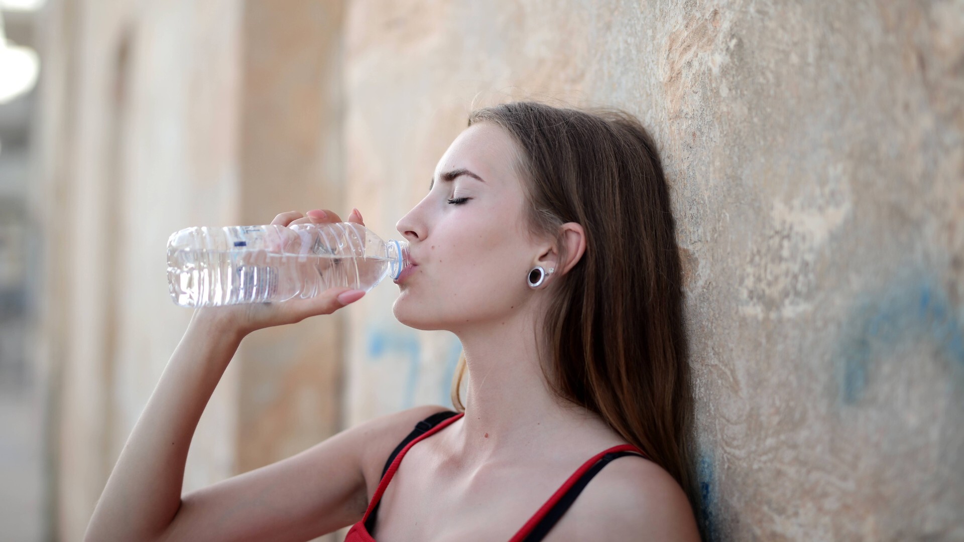 Eine Frau lehnt an einer Mauer und trinkt aus einer Waserflasche.