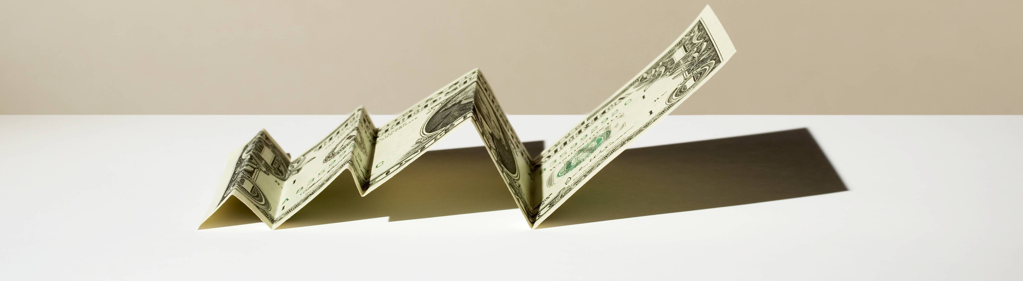 Ein gefalteter Dollar-Schein liegt auf einer Oberfläche. Der Dollar-Schein erinnert durch das Falten an einen Graphen, der ansteigt.