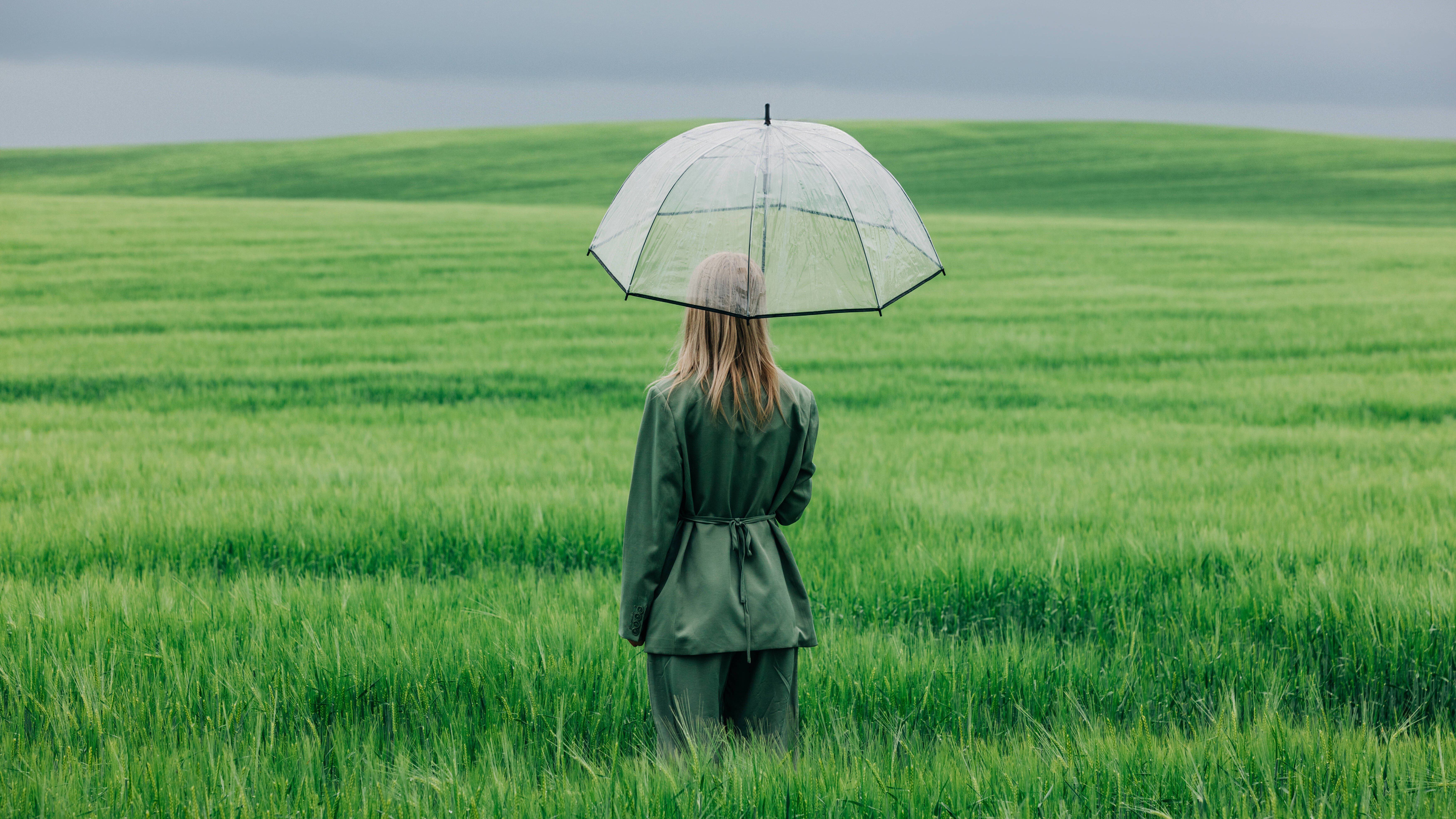 Modellfoto einer Frau, die mit einem durchsichtigen Regenschirm im Regen in einem grünen Weizenfeld steht