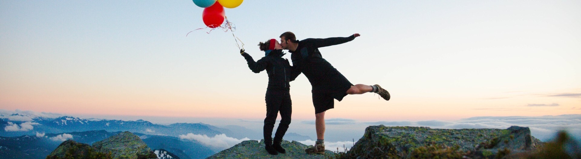 Eine Frau mit Luftballons in der Hand und ein Mann, der ihr einen Kuss gibt. Beide stehen auf der Spitze eines Berges.