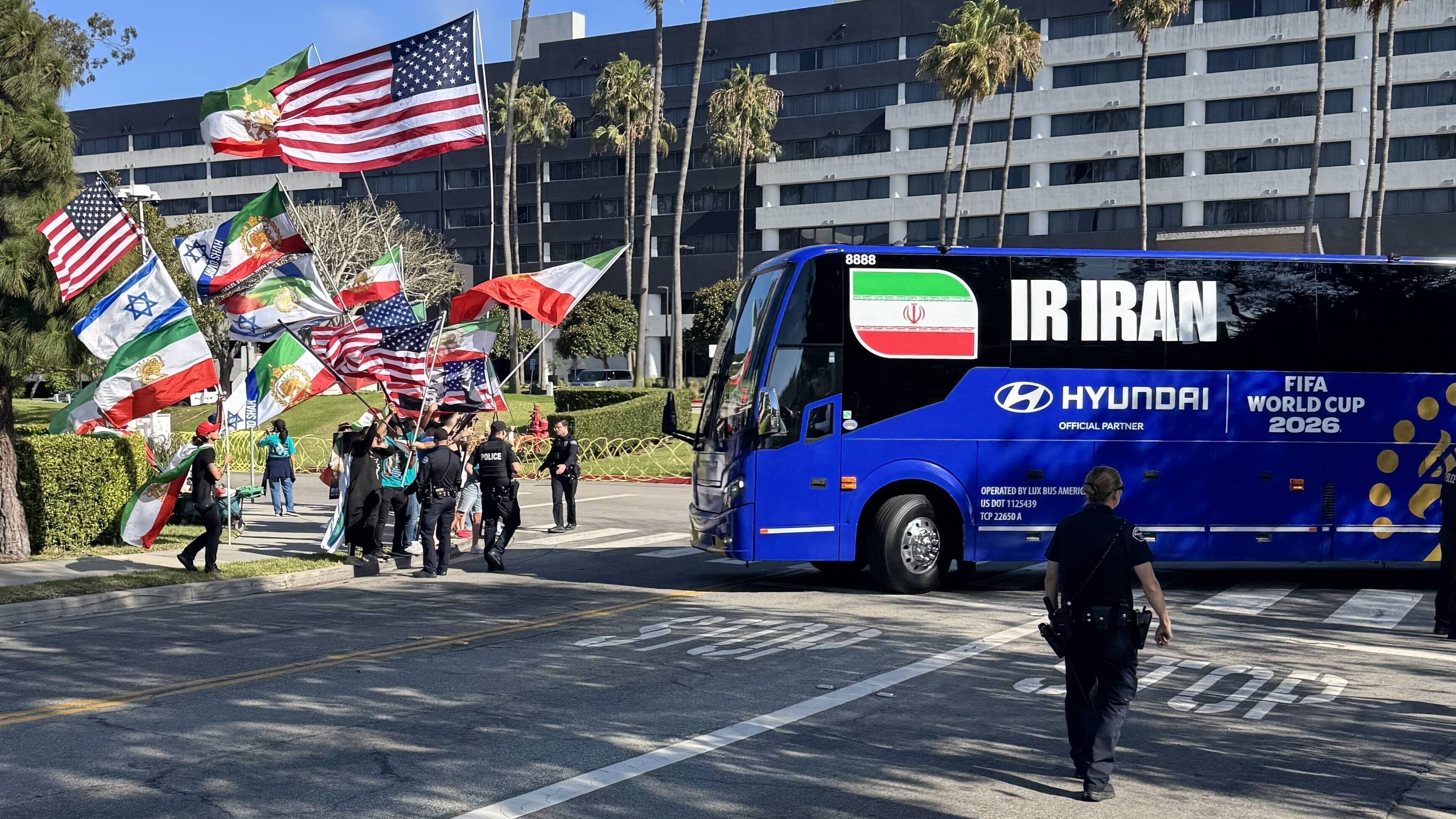 Protestierende schwenken die ehemalige iranische Flagge, die US-amerikanische und israelische Flaggen, als der Bus der Iranischen Fußballnationalmannschaft vor dem Hotel, in dem das Iranische Team untergebracht ist, wegfährt (Los Angeles, USA, 14.06.2026)