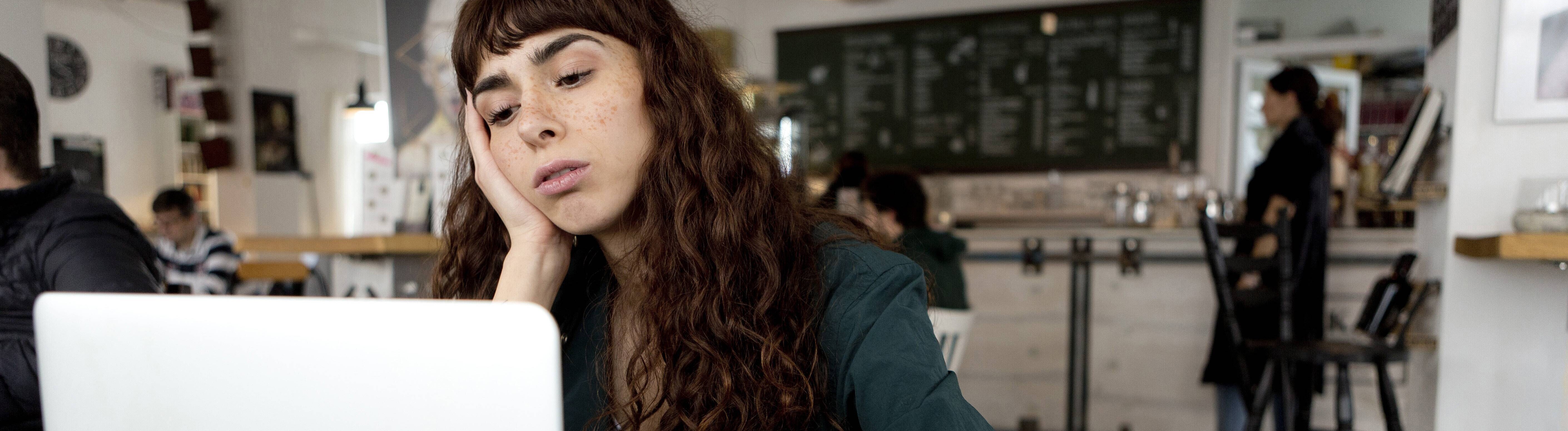 Eine Frau sitzt in einem Café vor einem aufgeklappten Laptop. Sie hat ihren Kopf auf einer Hand abgestützt und schaut gelangweilt auf den Bildschirm.
