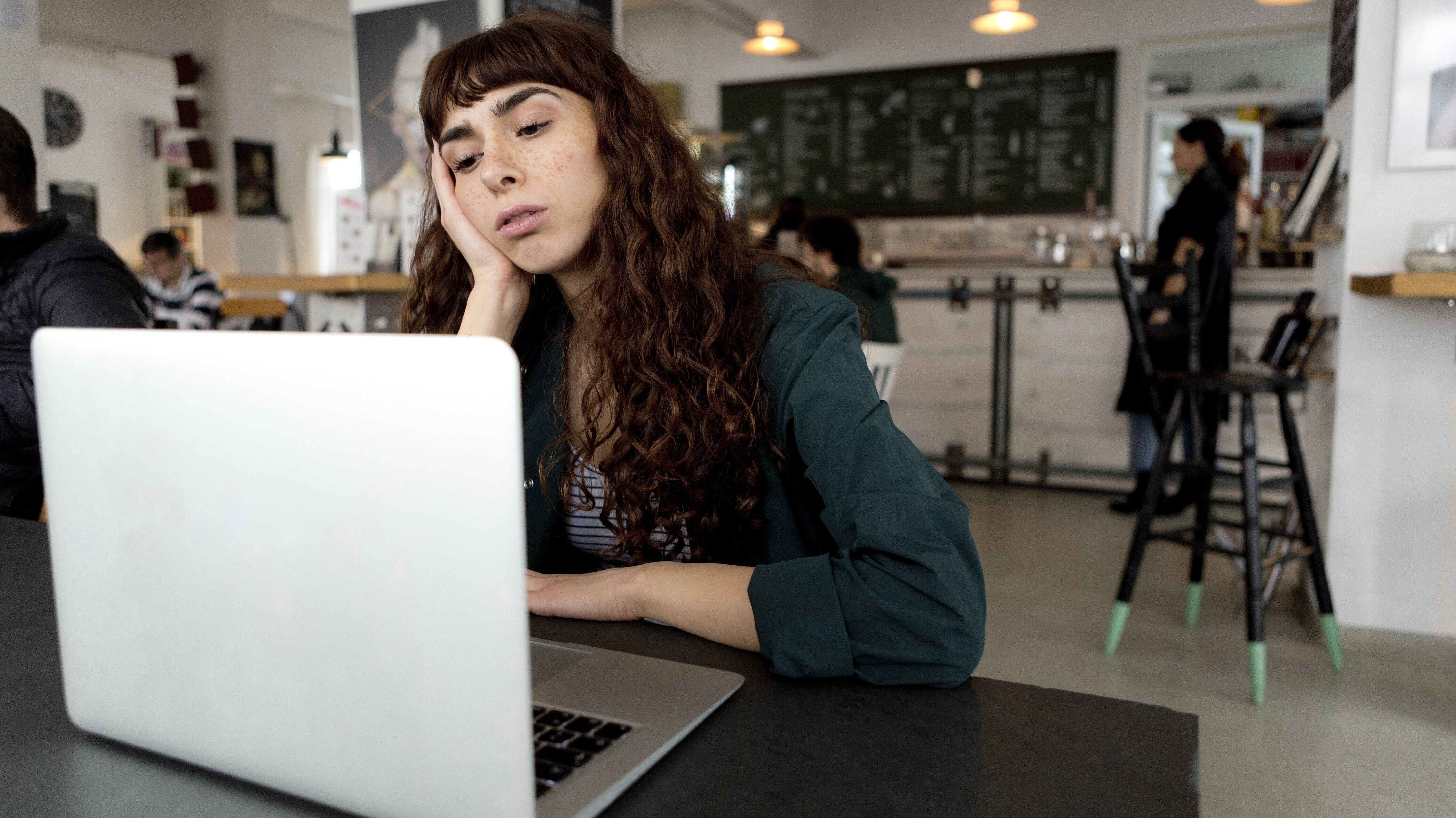 Eine Frau sitzt in einem Café vor einem aufgeklappten Laptop. Sie hat ihren Kopf auf einer Hand abgestützt und schaut gelangweilt auf den Bildschirm.