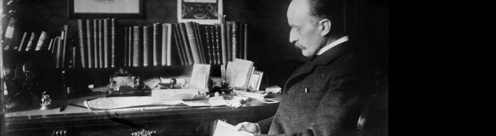 Das Schwarz-Weiß-Foto zeigt den deutschen Physiker Max Planck in seinem Arbeitszimmer während er an seinem Schreibtisch sitzt und in ein Buch schaut.