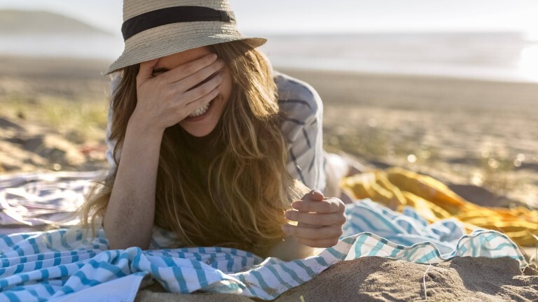 Eine Frau liegt mit dem Rücken zum Meer am Strand. Die Frau trägt einen Sonnenhut und eine Hemd und liegt auf ihrem Bauch auf einem Tuch. Sie hält ihre Hand vor ihre Augen und lacht.